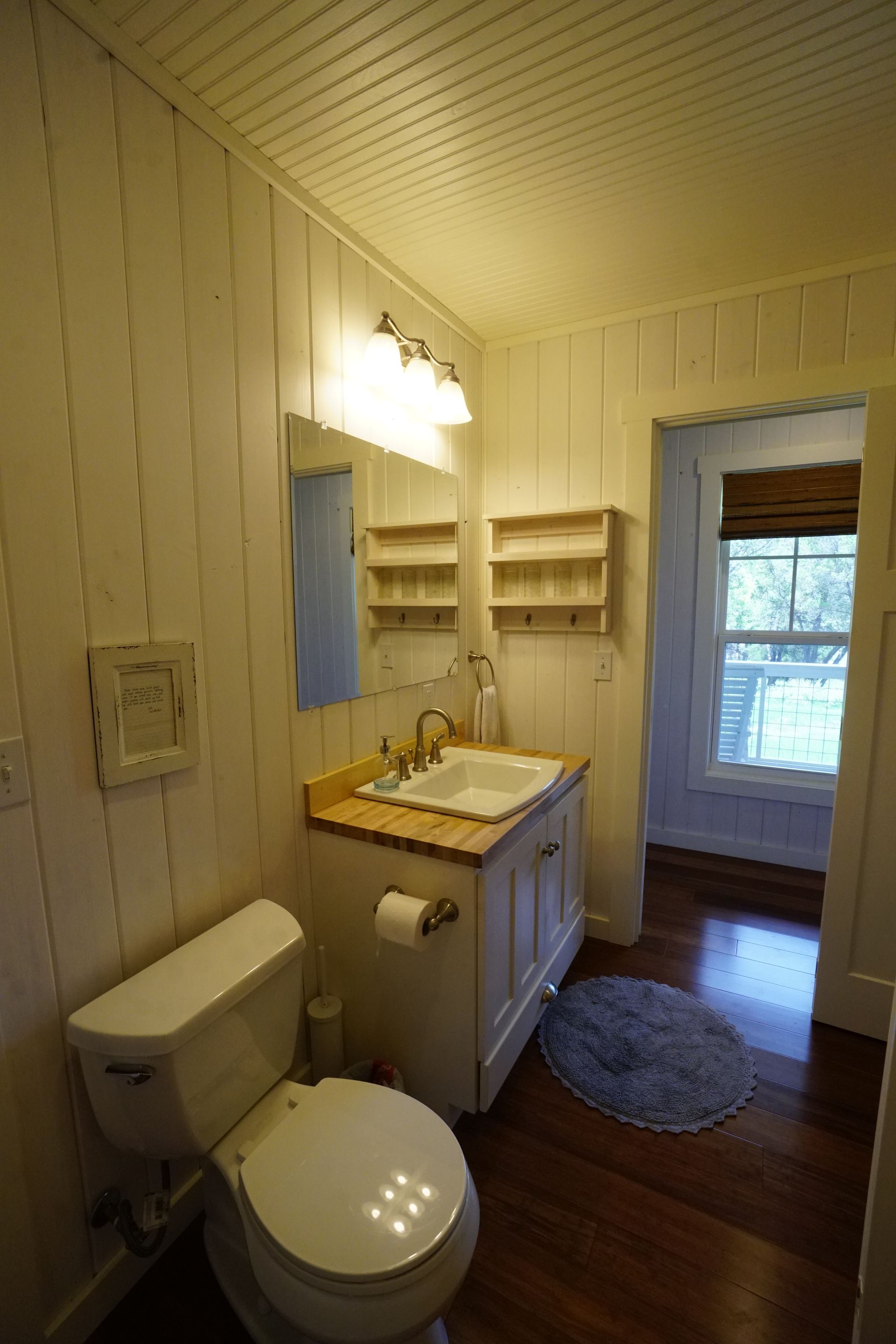 Small white-paneled bathroom with a toilet, sink, mirror, and open doorway to another room with window.
