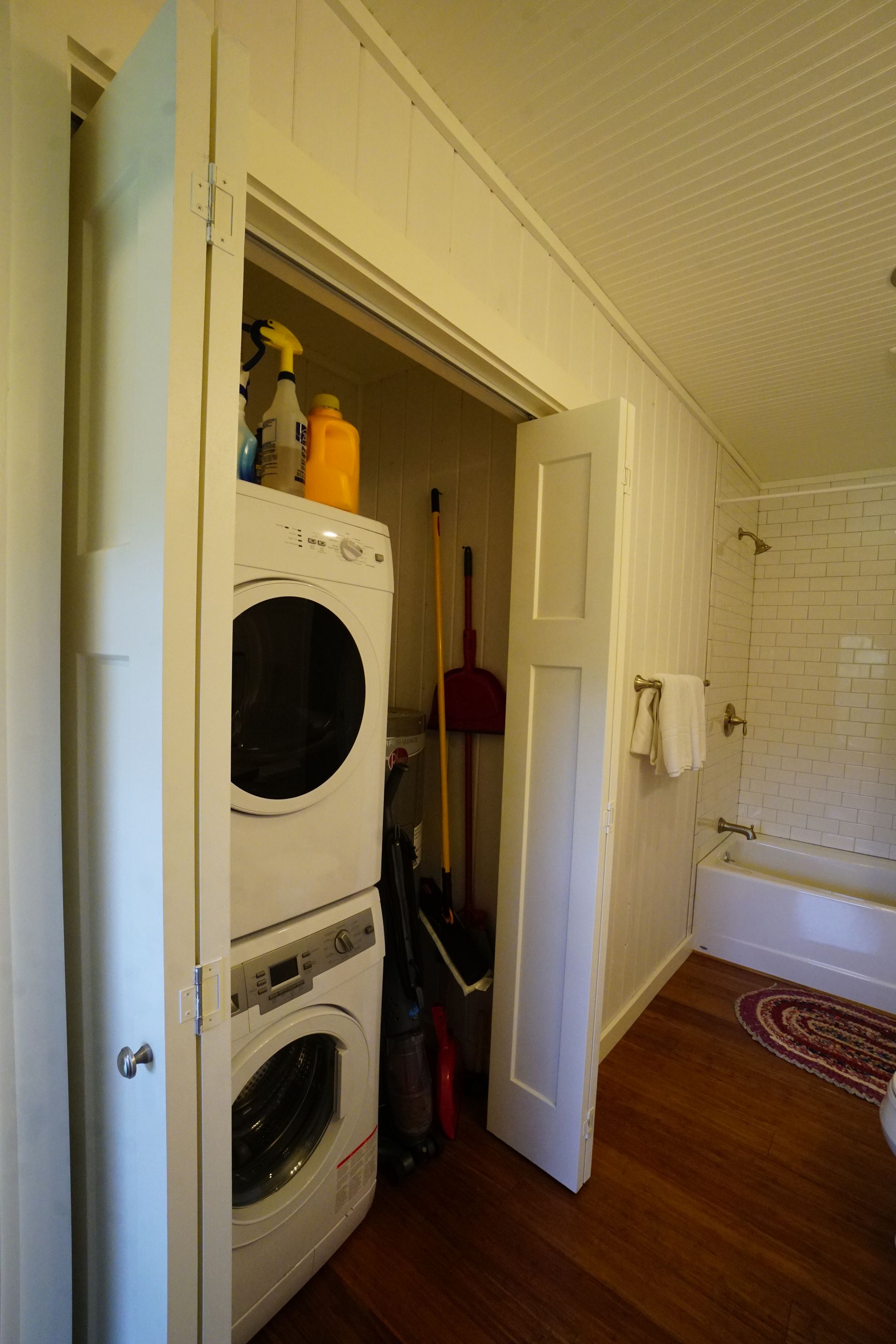 Stacked washer/dryer inside a closet with cleaning supplies. Bathroom with tub visible. Wooden floor.