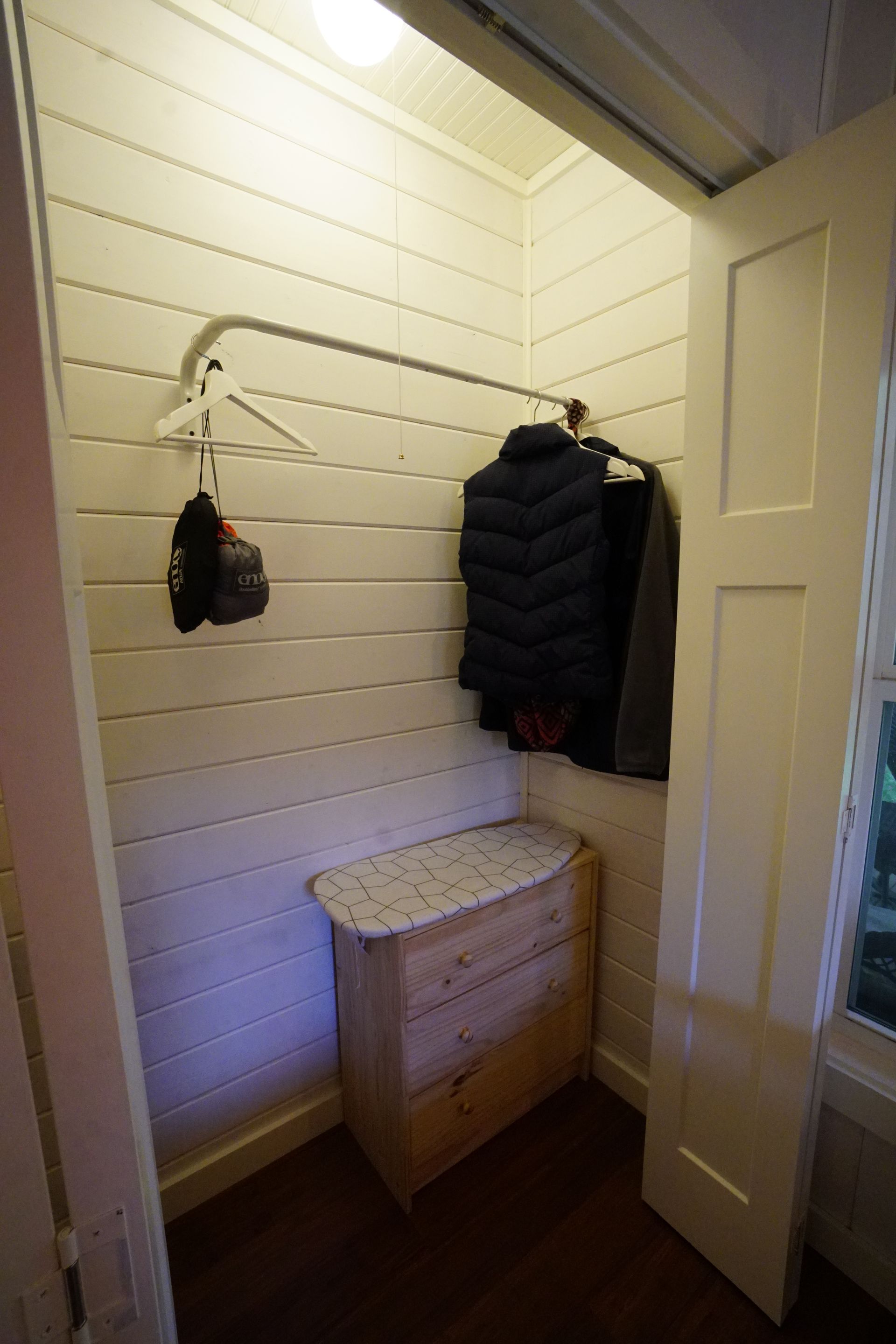 Small closet with clothes hanging on a rod, a wooden bench, and white paneled walls.