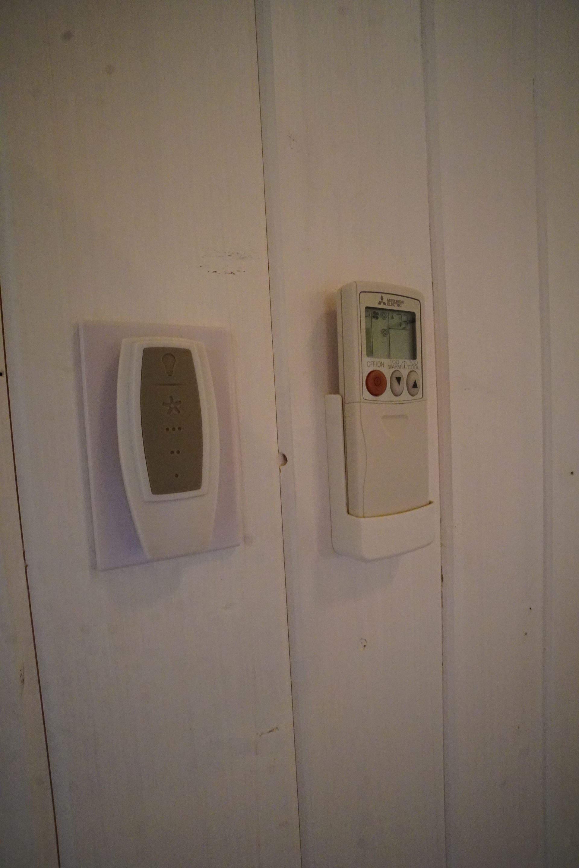 Two white wall-mounted climate control devices. One rectangular with a small screen, the other has a rounded design.