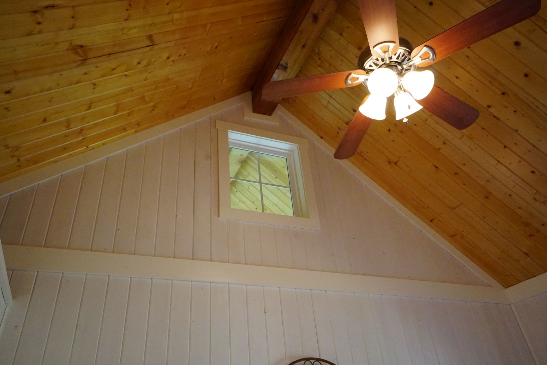 Interior of a room with a wood ceiling, paneled walls, a small window, and a ceiling fan.