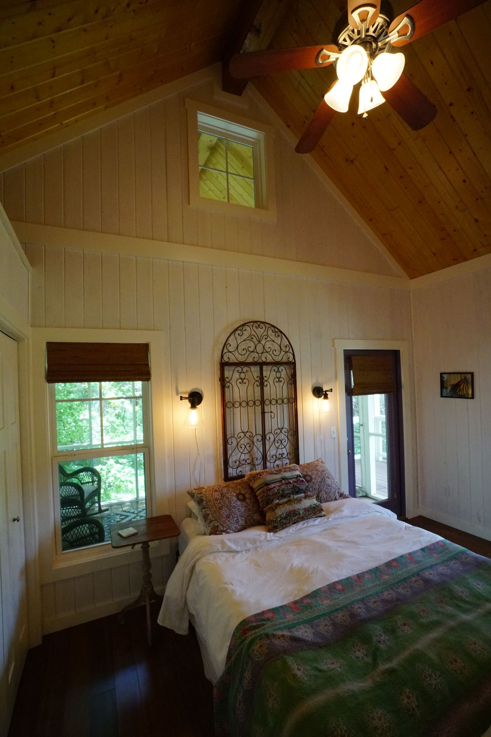 Bedroom with white paneling, high wood ceiling, bed, windows, and door.