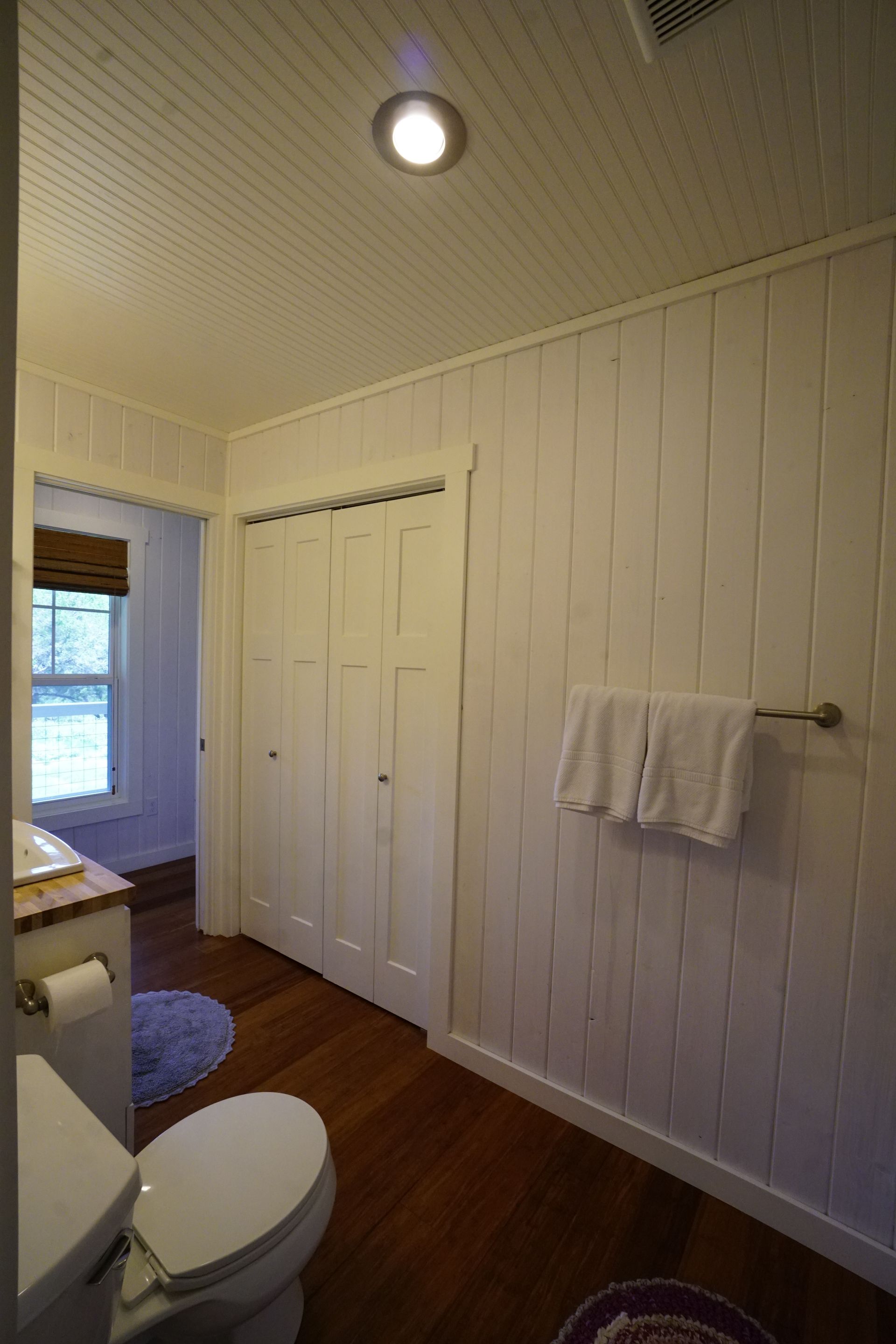 Bathroom with white panel walls, closet, toilet, and towels on a rack. Brown wooden floor.