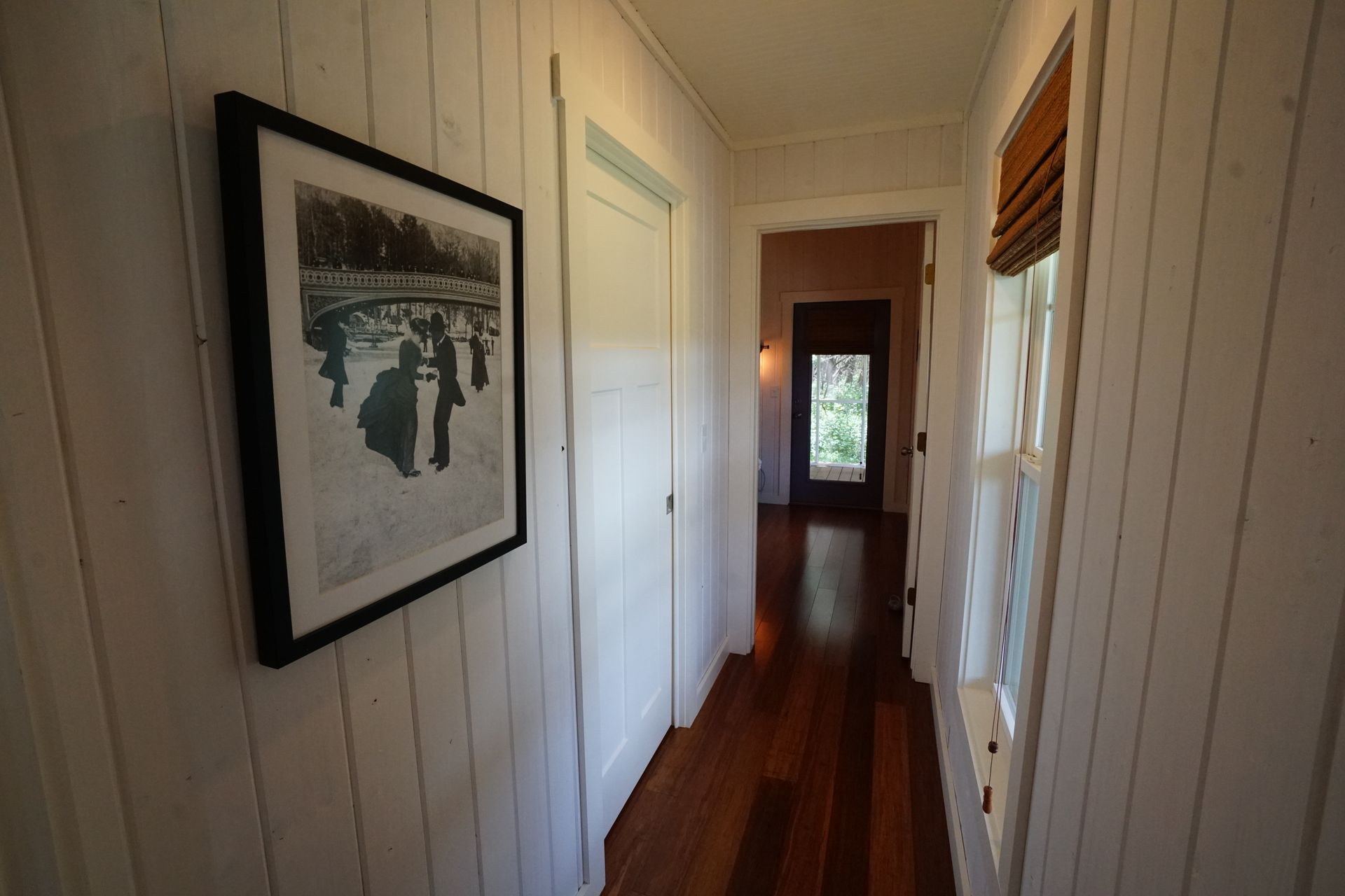 Hallway with white paneled walls, wood floor, artwork, and a window with a shade.