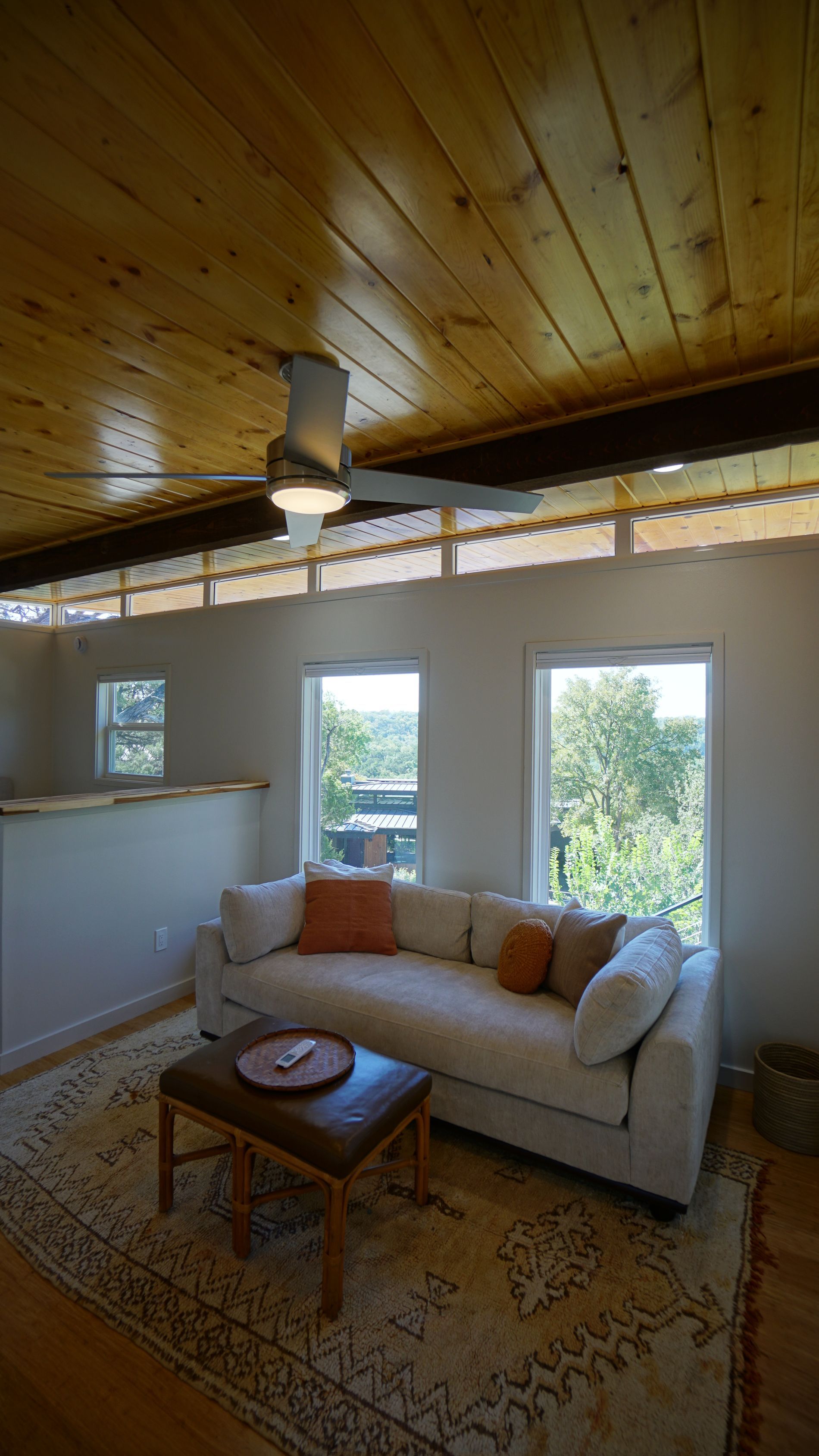 Living room with white sofa, rug, and wooden ceiling. Windows overlook green trees.