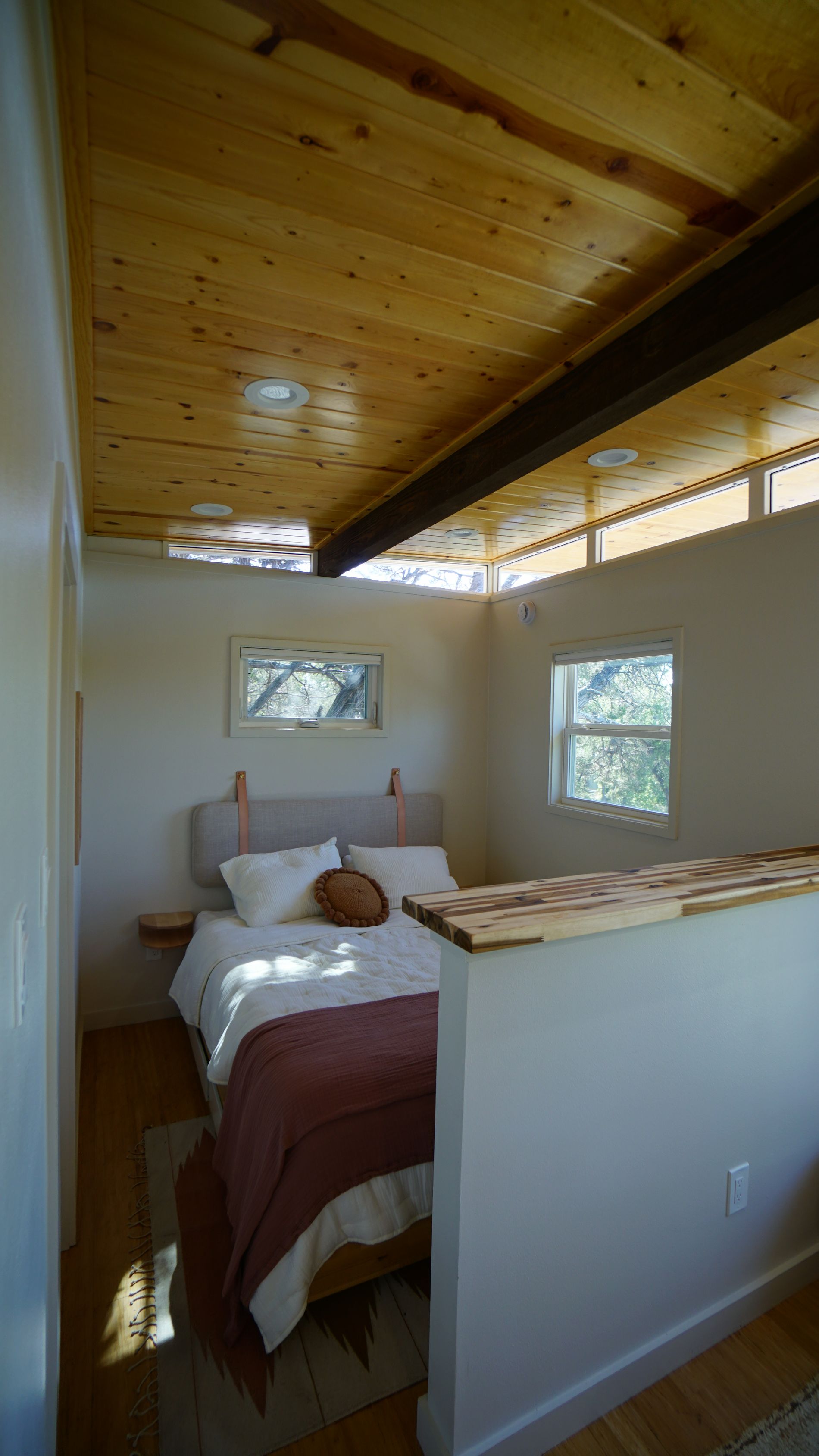 Bedroom with wood ceiling, bed, windows, and partial wall.  Natural light and neutral colors.