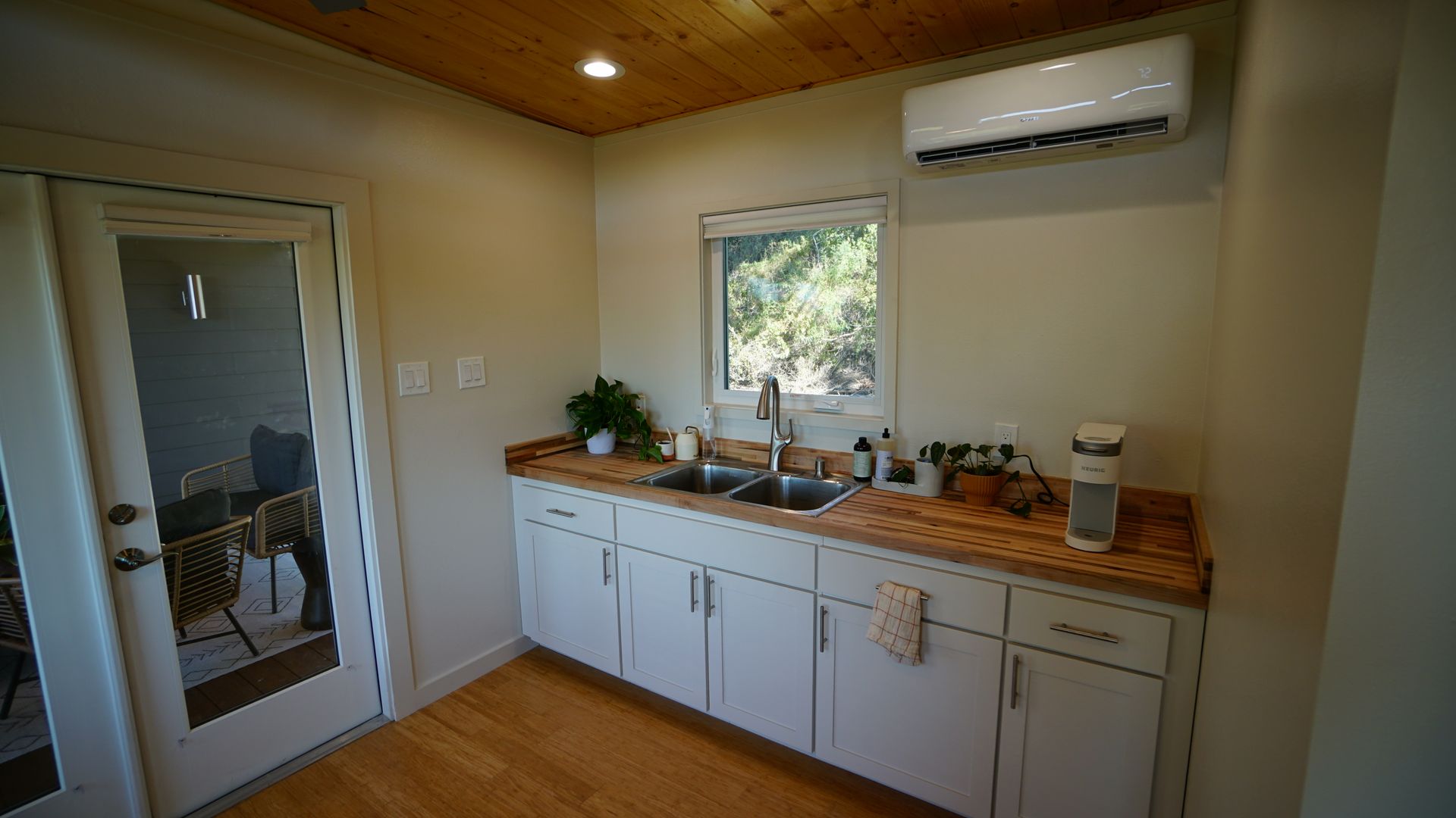 Small kitchen with white cabinets, wooden countertop, window above sink, air conditioning unit, and glass door.