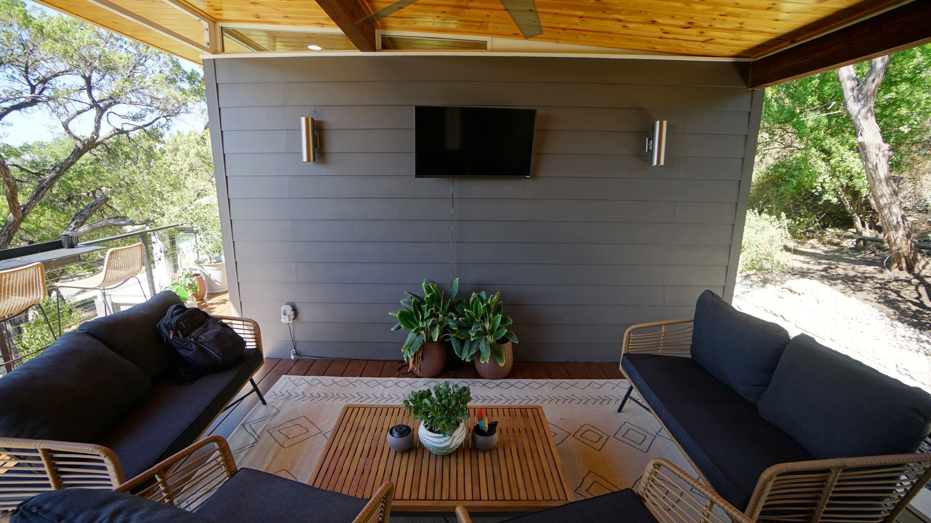 Outdoor living space with gray wall, TV, couches, coffee table, rug, plants, and trees.