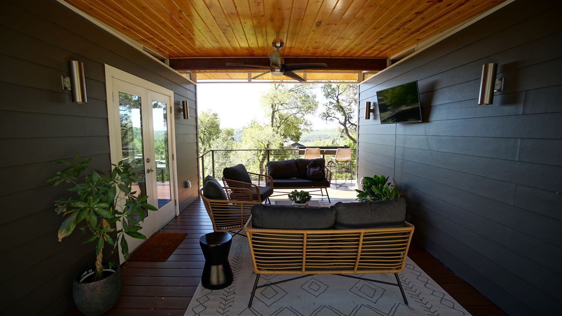 Outdoor patio with seating, trees, and TV. Grey walls, wooden ceiling, and rug.