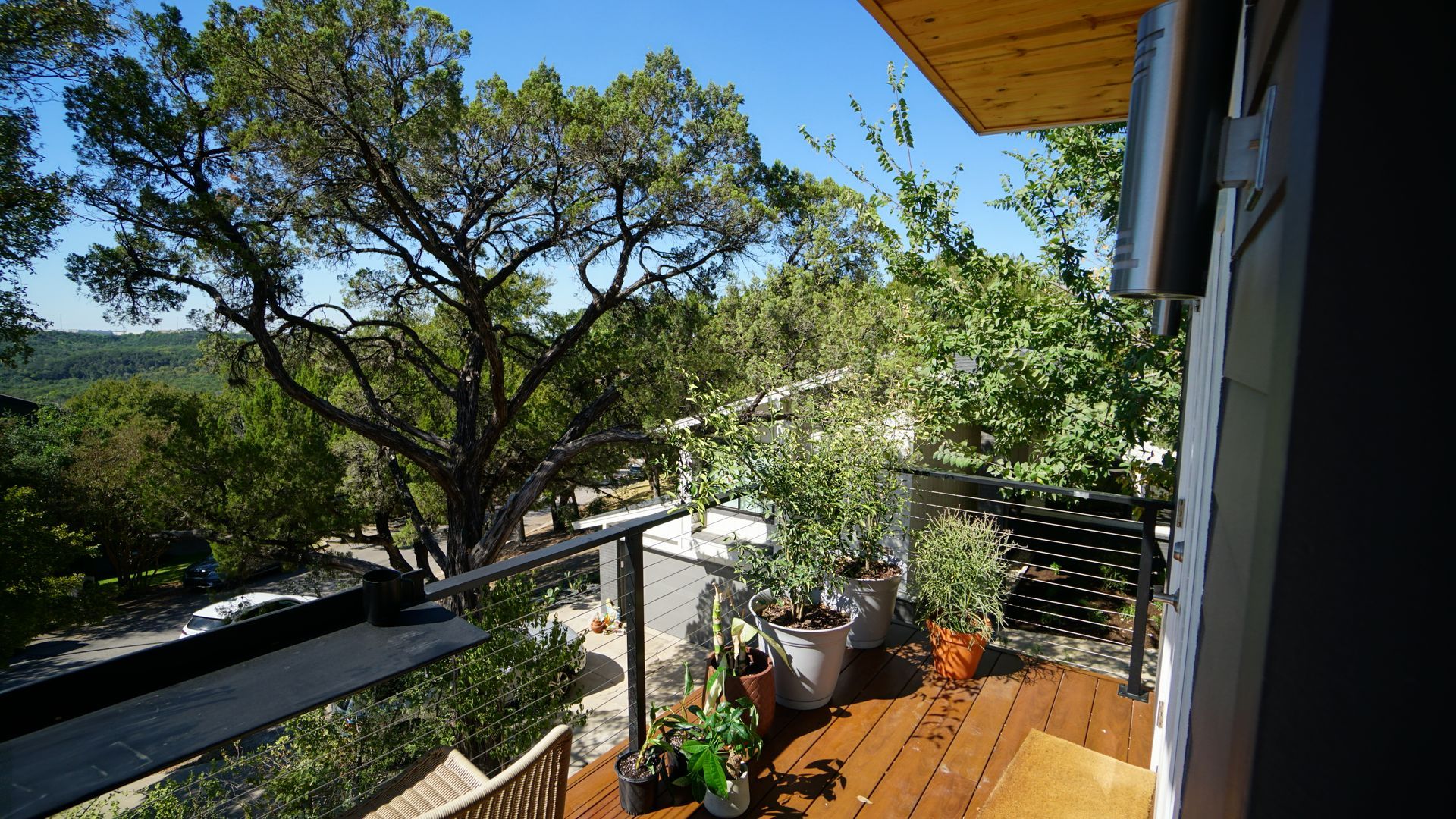 Balcony with glass railing, potted plants, and wooden deck overlooking a tree-filled landscape on a sunny day.