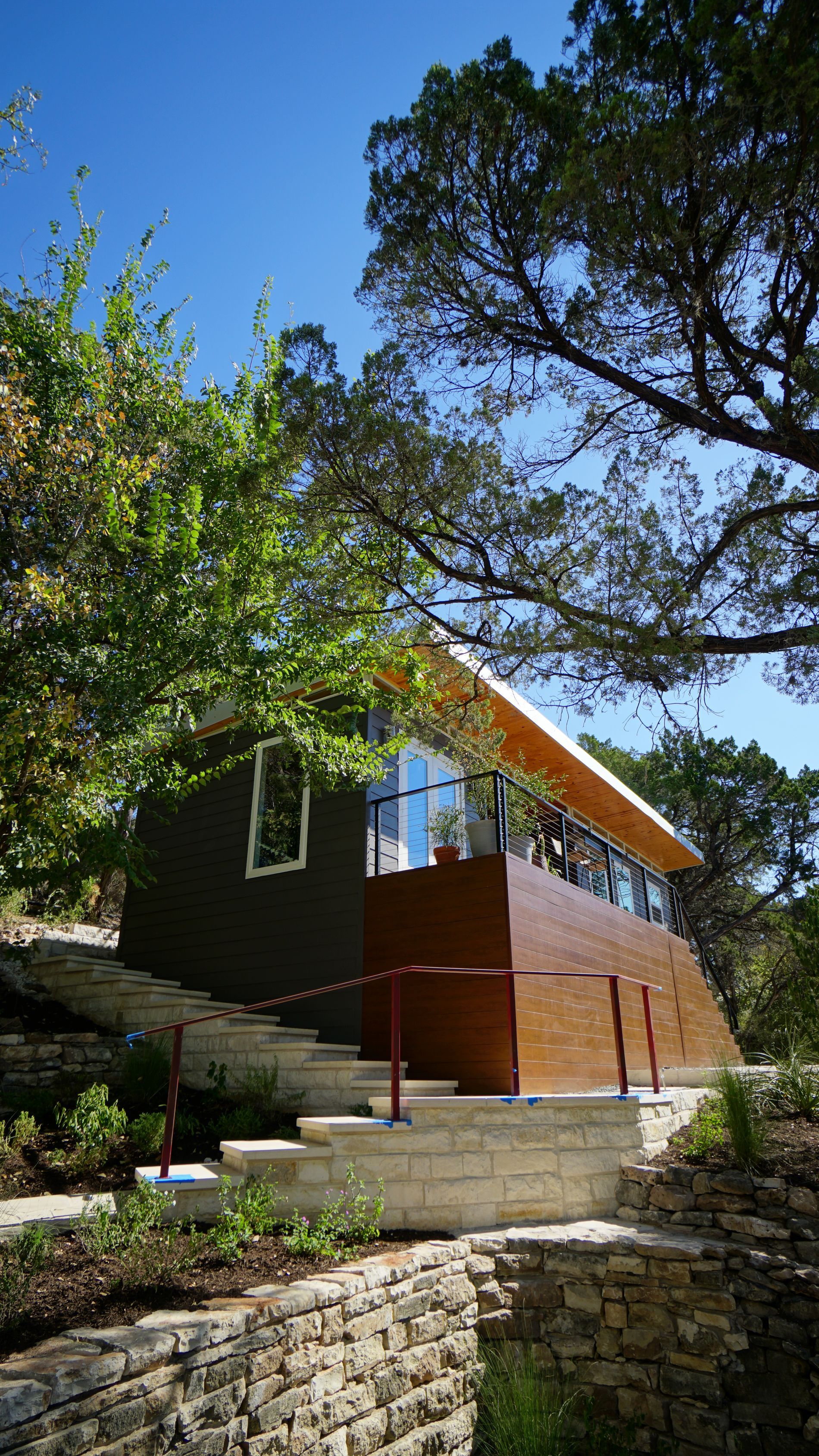 Modern house with brown and dark gray exterior, stairs, and surrounding trees against a blue sky.