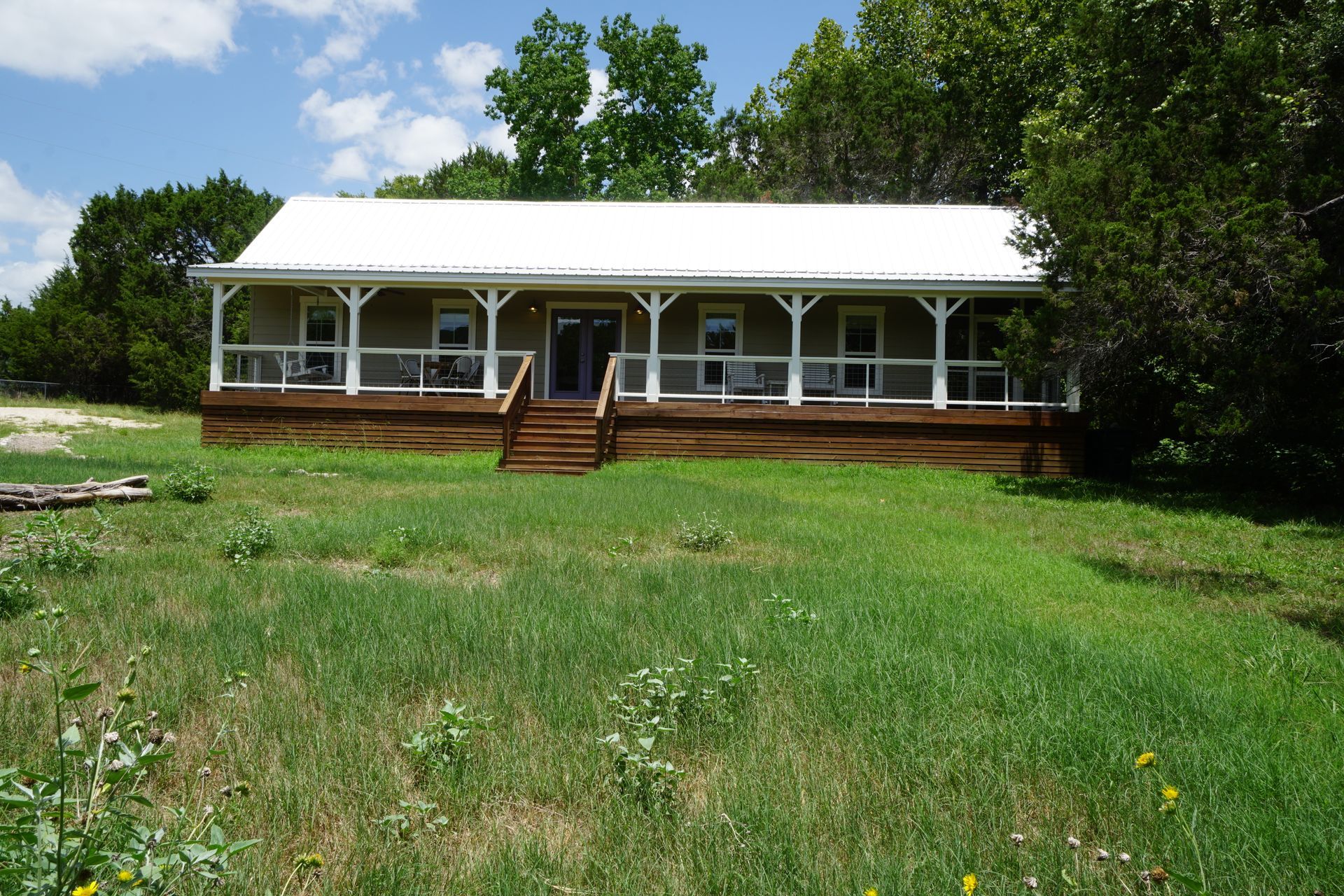 Tan house with white porch and roof, wooden steps, surrounded by green grass and trees.