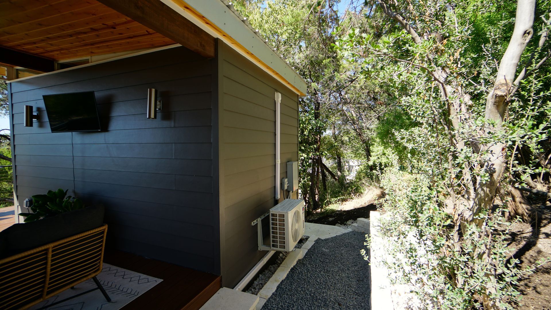 Exterior view of a dark grey cabin with a covered porch and gravel pathway leading into a wooded area.