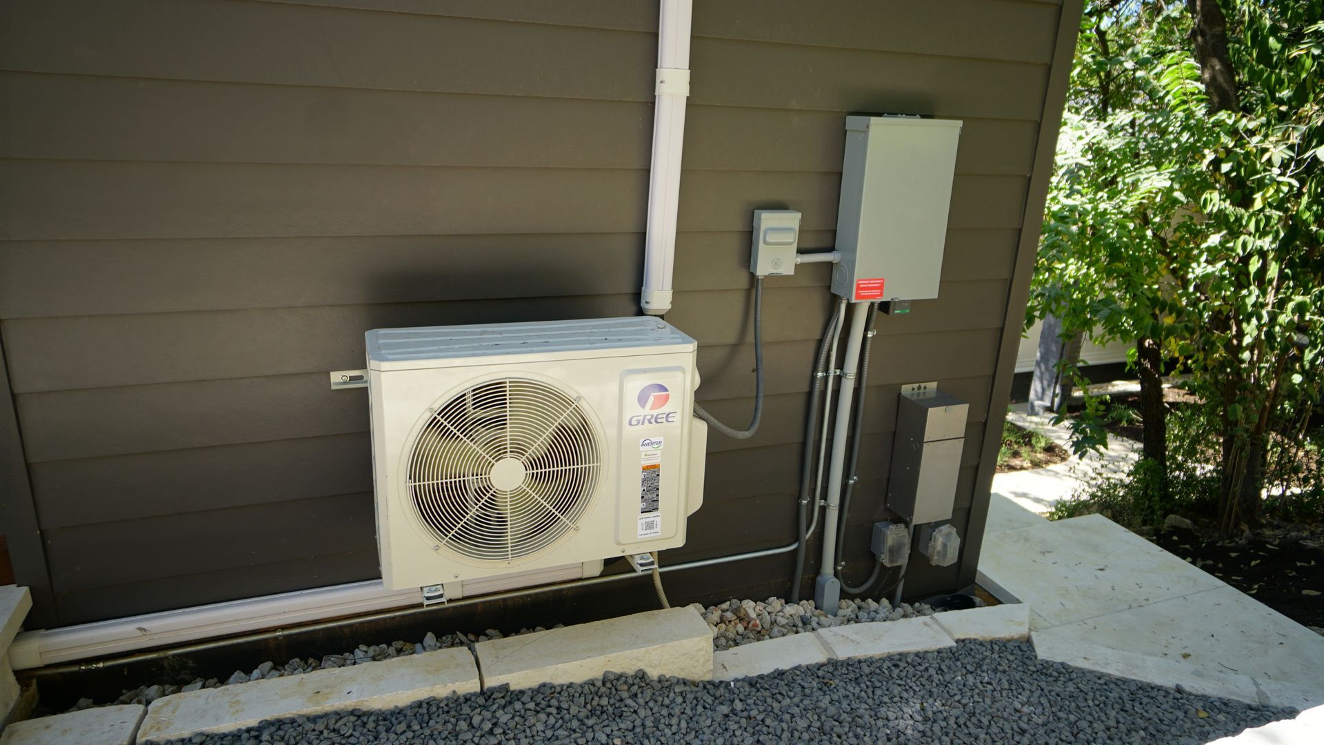 Outdoor HVAC unit and electrical box mounted on a brown wall with gravel ground.
