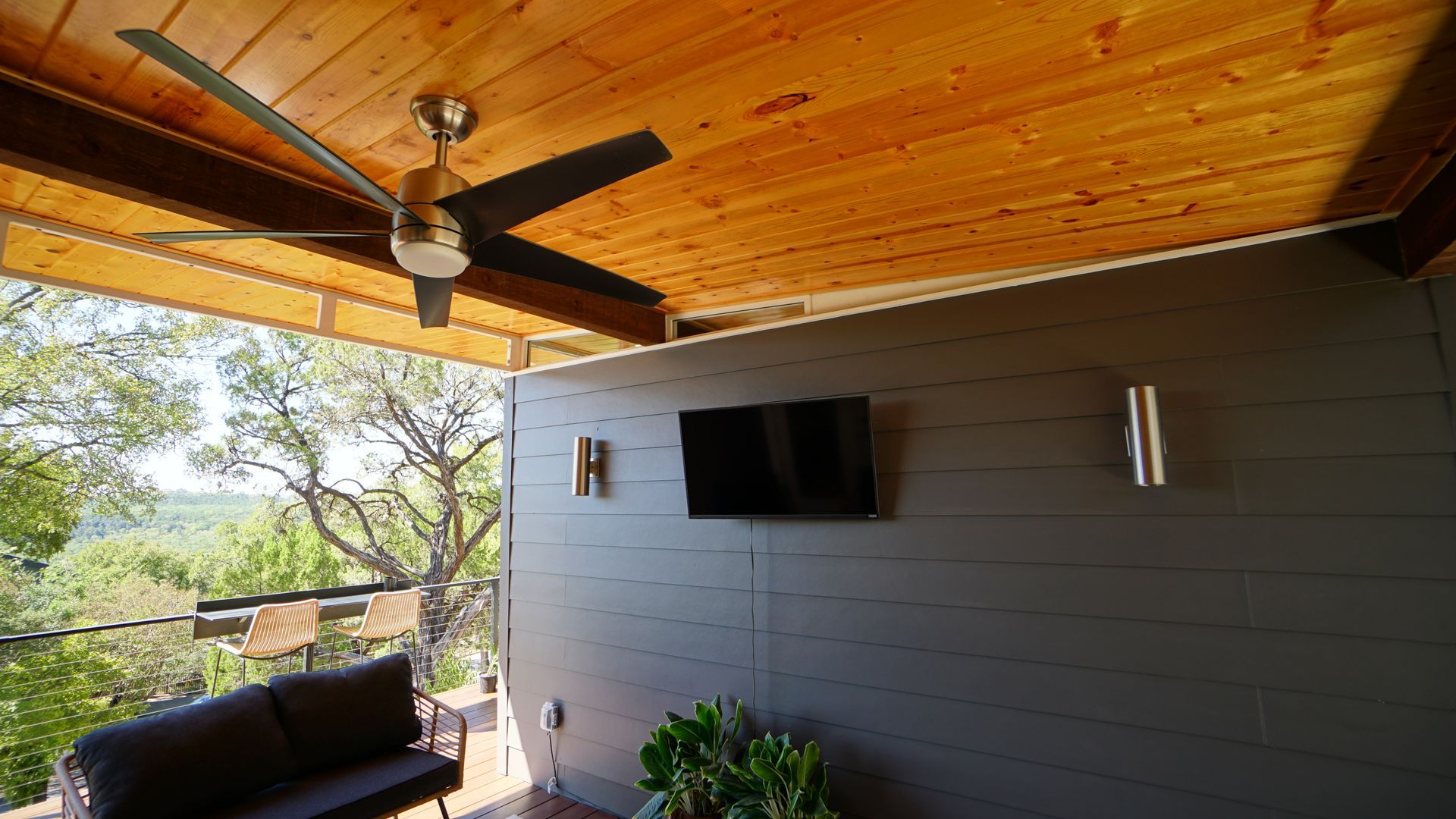 Outdoor deck with a ceiling fan, TV, and gray wall overlooking a green landscape.