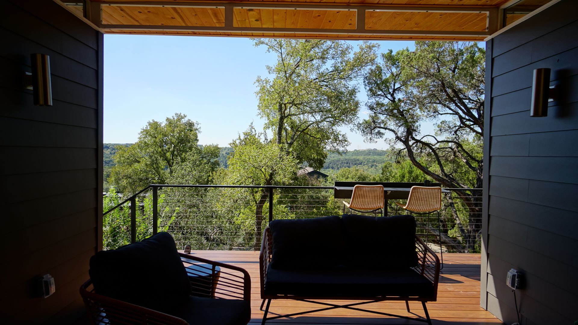 View from an open doorway onto a deck with seating, overlooking a forest under a blue sky.