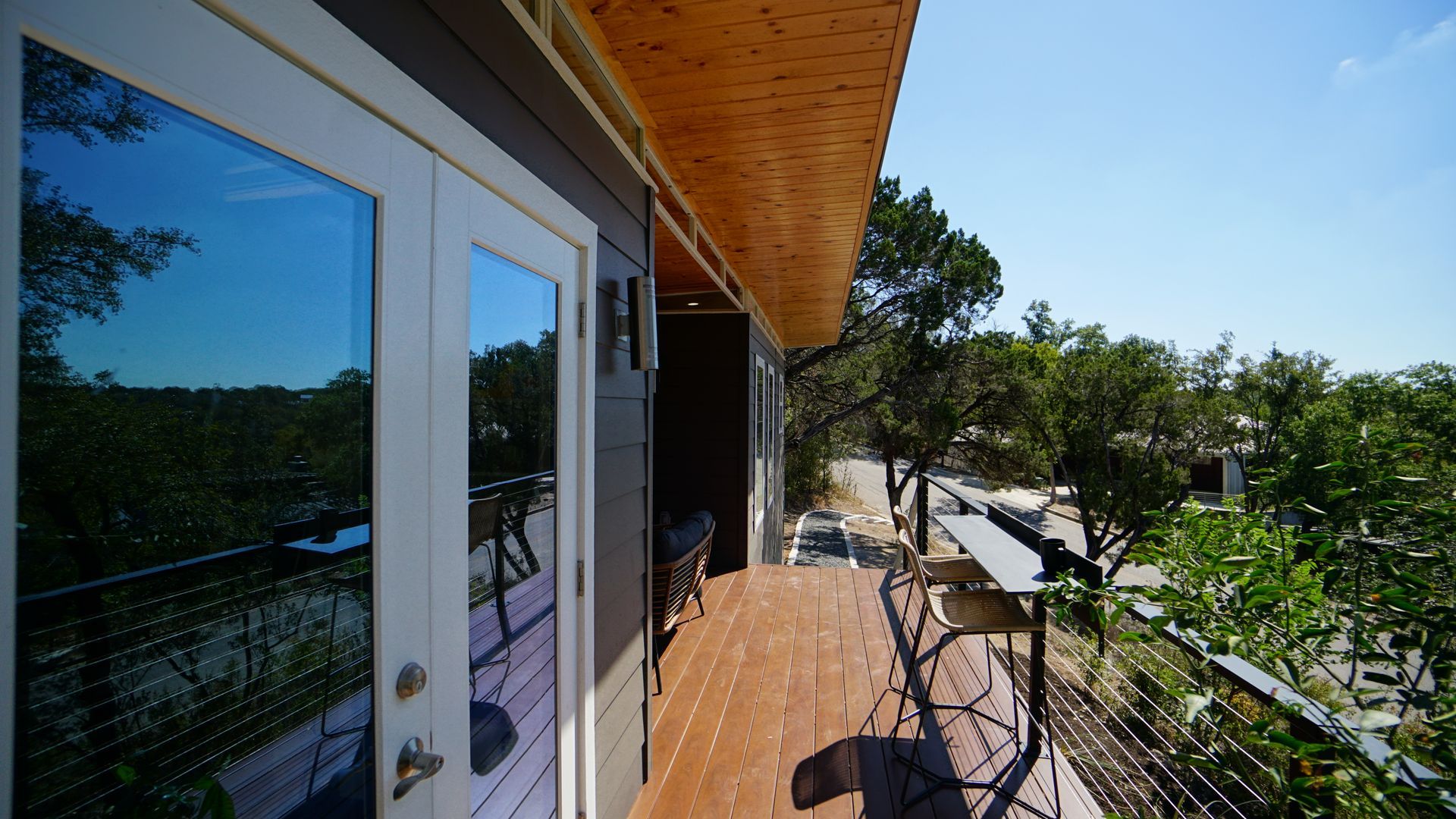 Exterior view of a modern home with a wooden deck and a glass door overlooking a lush landscape on a sunny day.