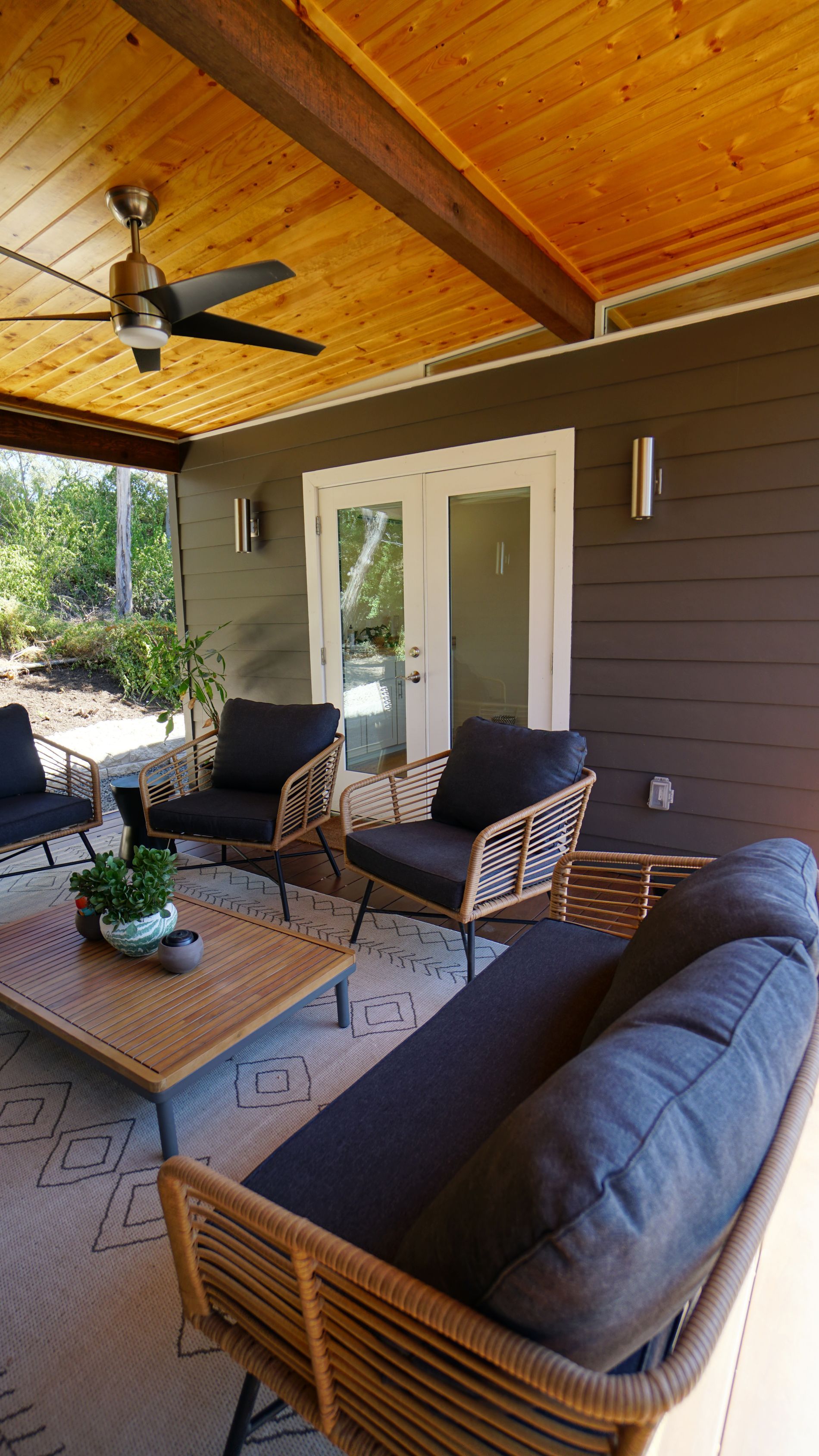 Covered patio with wicker furniture and black cushions, wood ceiling, and double glass doors.