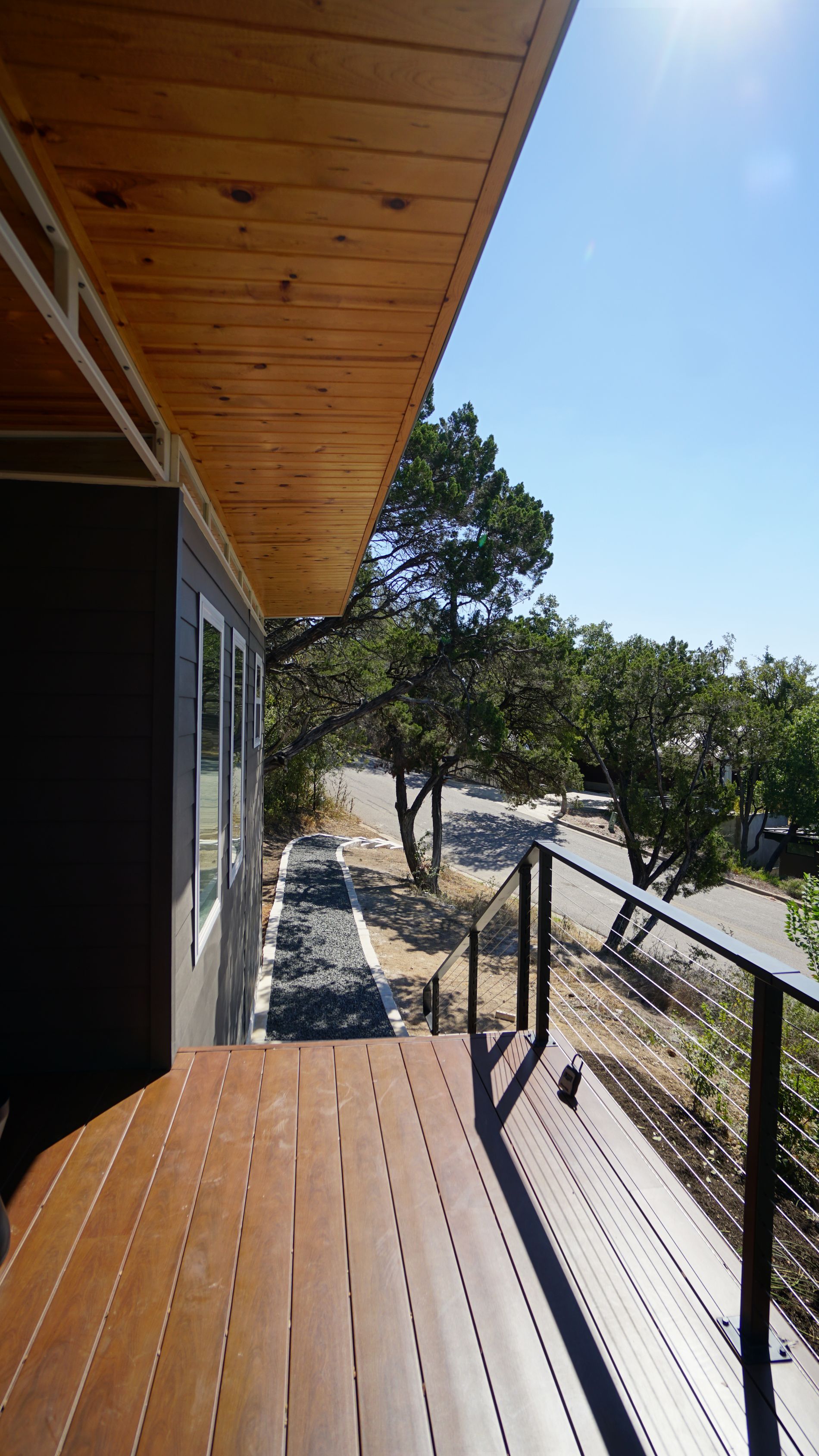 Exterior view from a wooden deck with railing, trees, and road. Brown wood table, blue sky.