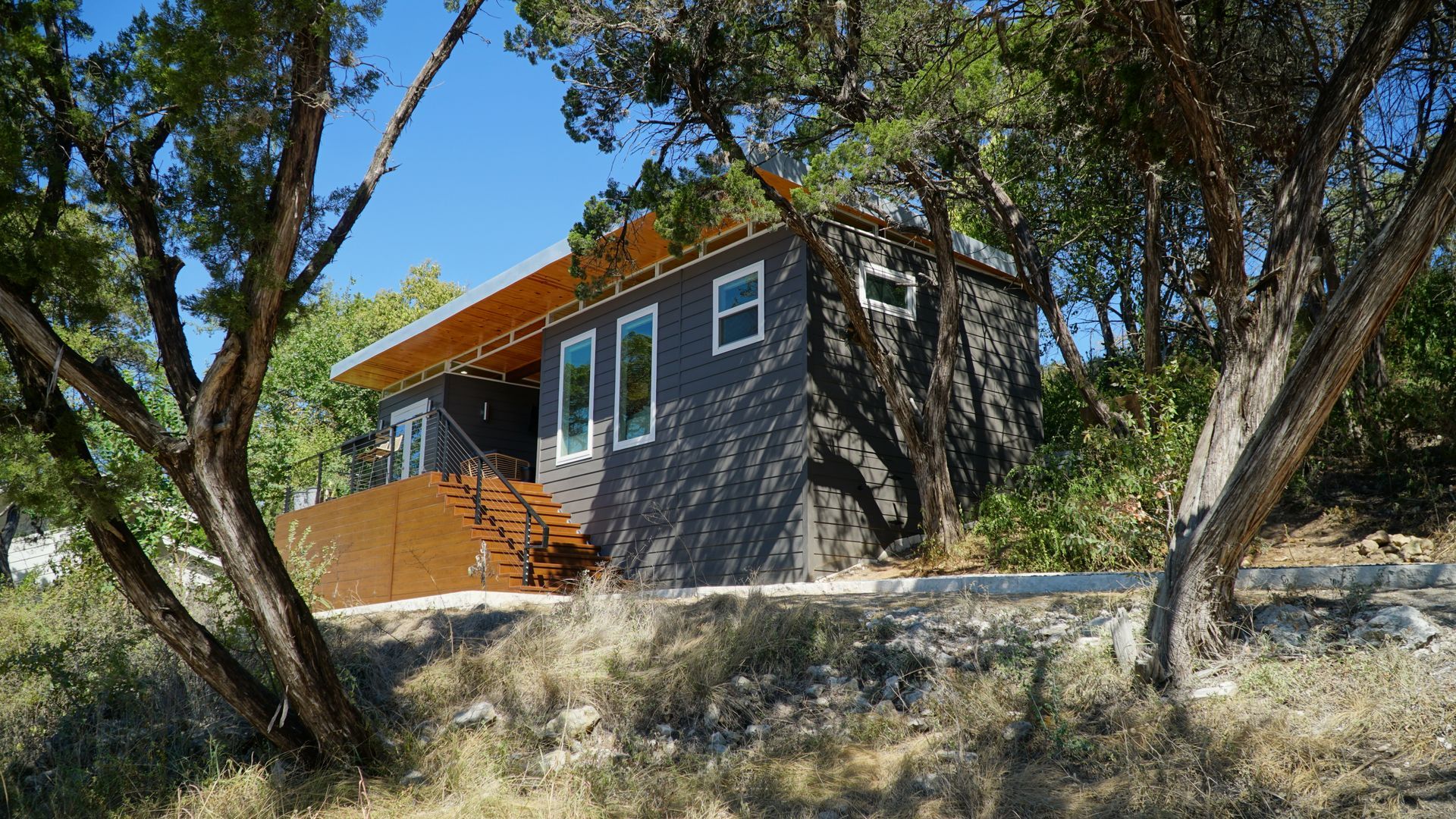 Modern cabin with dark gray siding, wooden deck, and surrounding trees.