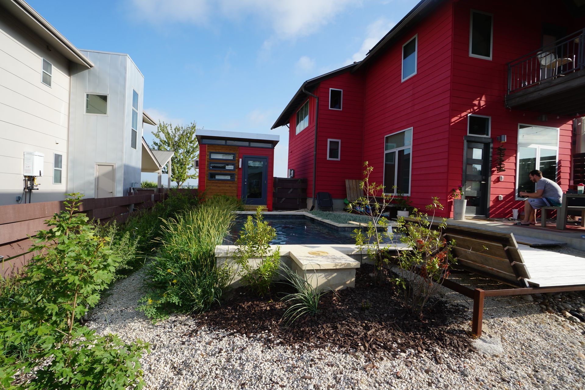 Red house with small pool, wooden structure, landscaping, and person sitting outside.