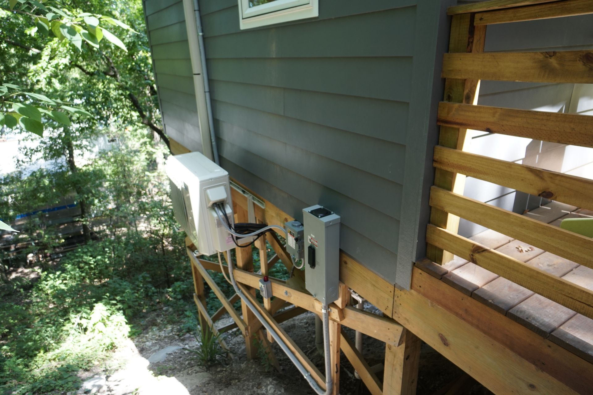 Exterior view of a gray-sided building with deck, electrical equipment mounted on the side, surrounded by greenery.