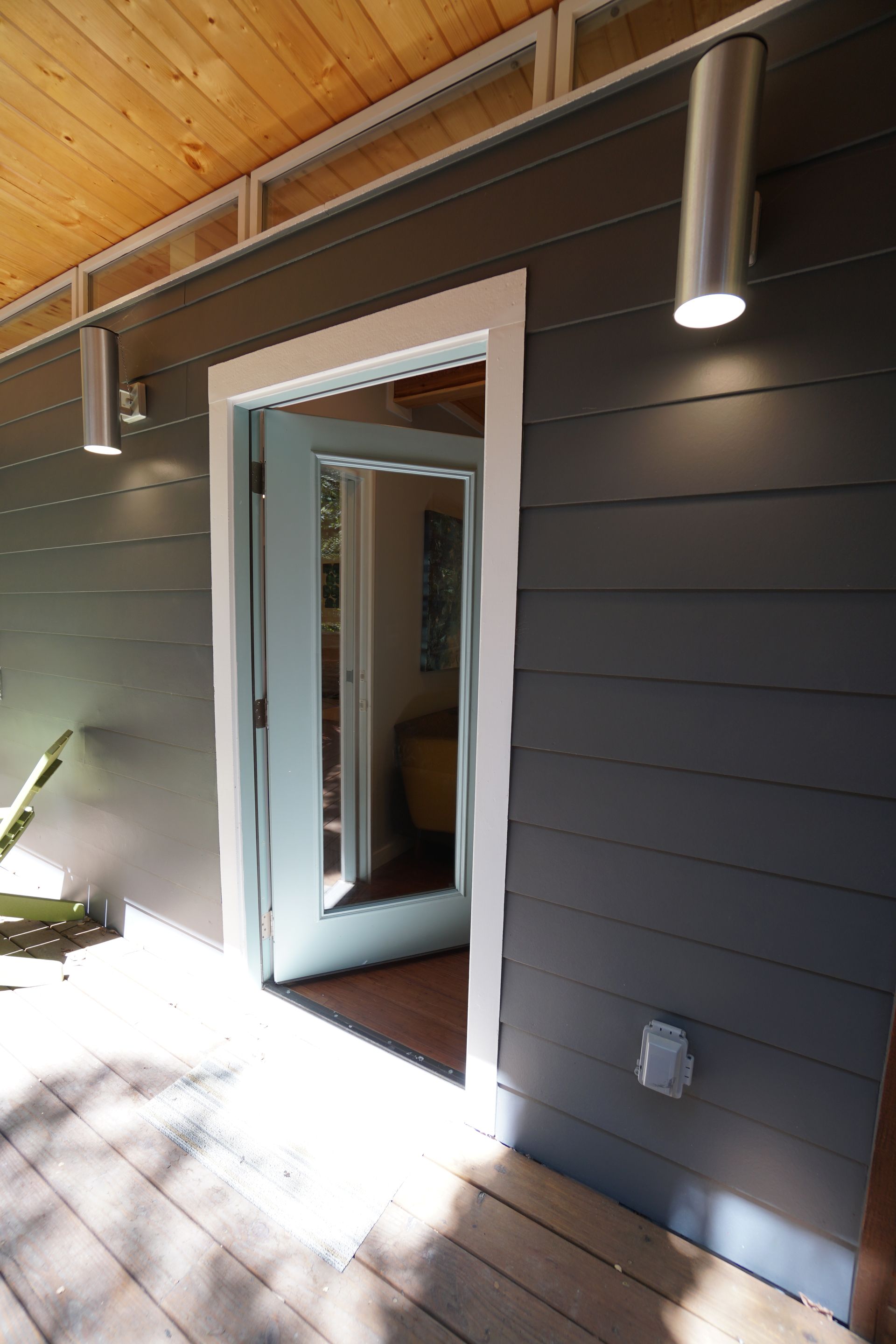 Exterior view of a door with white trim, gray siding, wooden deck, and modern light fixtures.