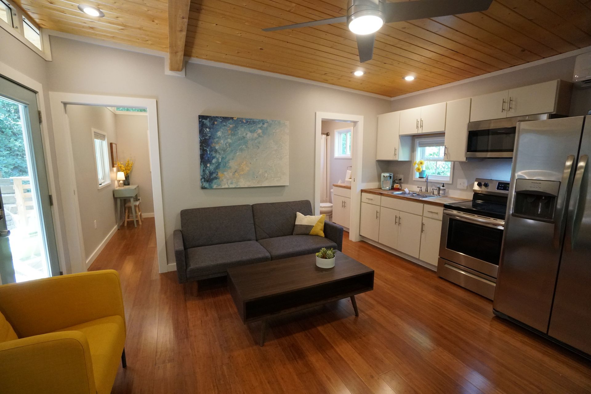 Cozy living space with kitchen. Grey sofa, yellow chair, wooden floors and ceiling, stainless steel appliances.