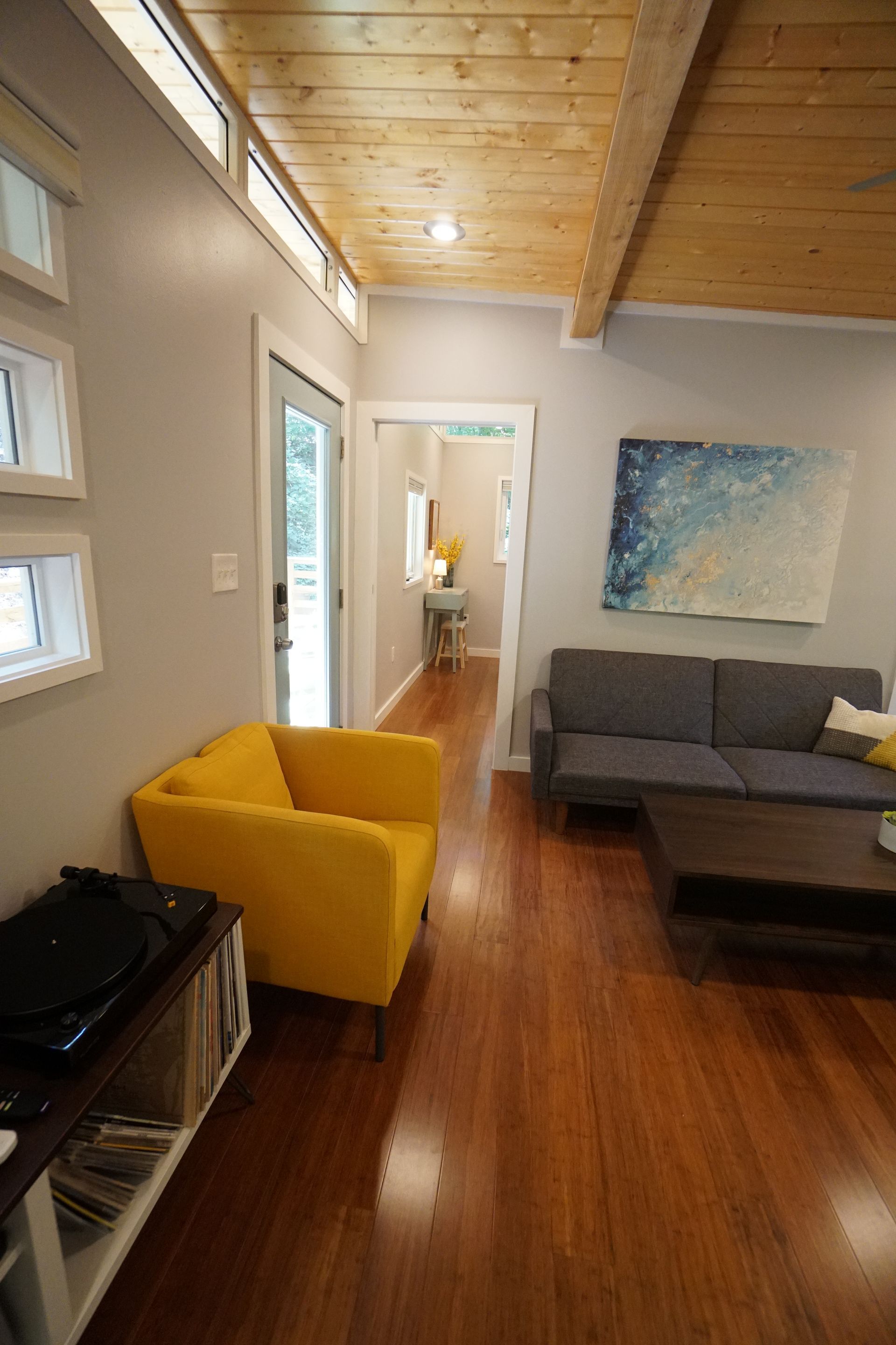 Living room with wood floors, yellow armchair, gray sofa, art, and a view into a hallway.