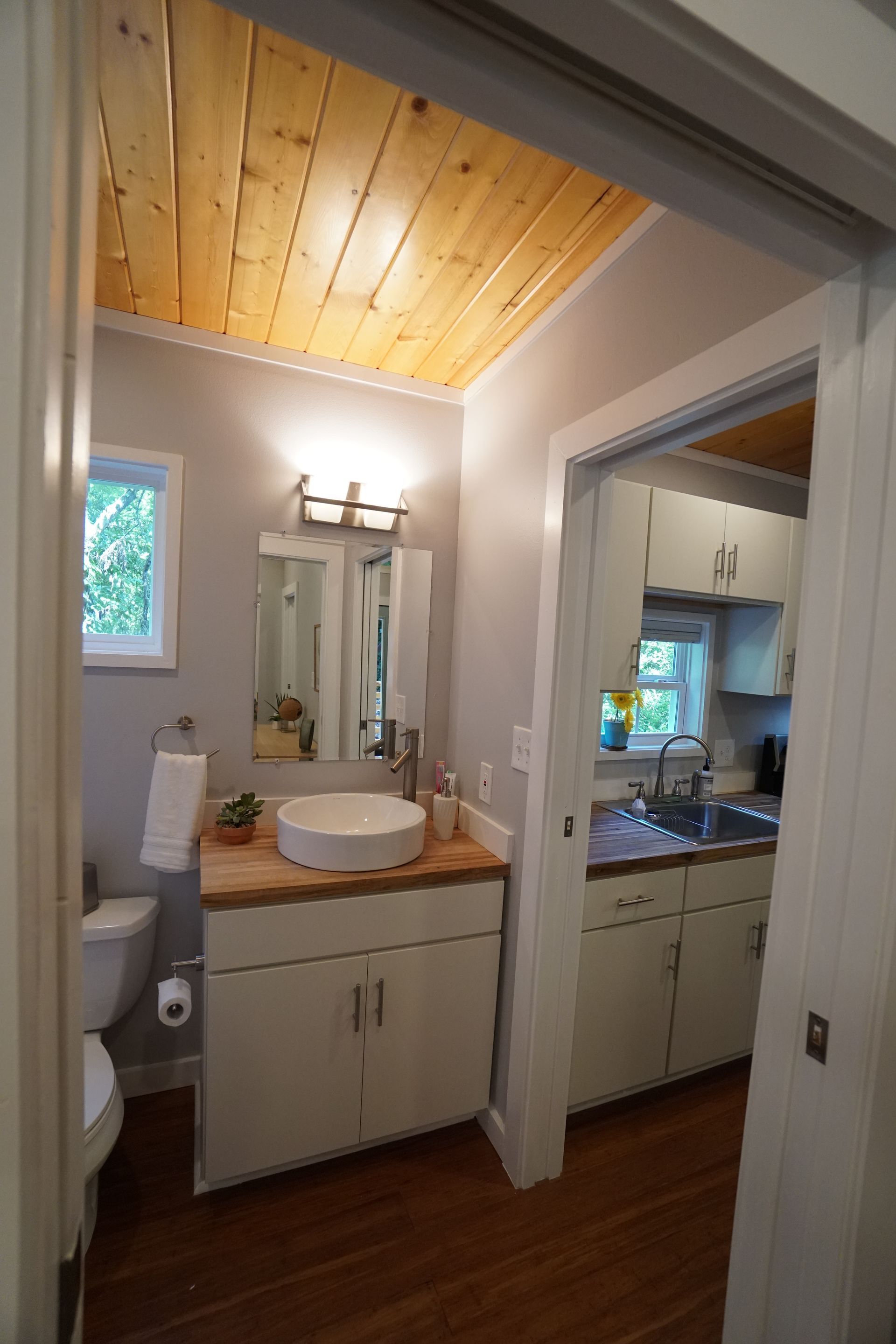 Bathroom interior with white vanity, toilet, and wooden ceiling, open to kitchen.