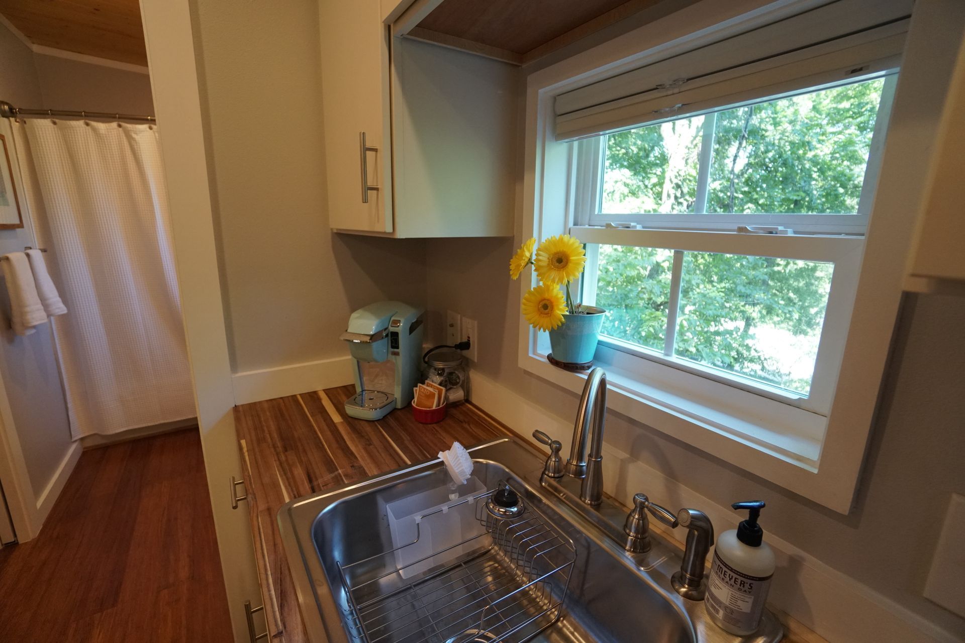 Kitchen with sink, cabinets, window, yellow flowers in a pot, and a K-cup coffee maker.