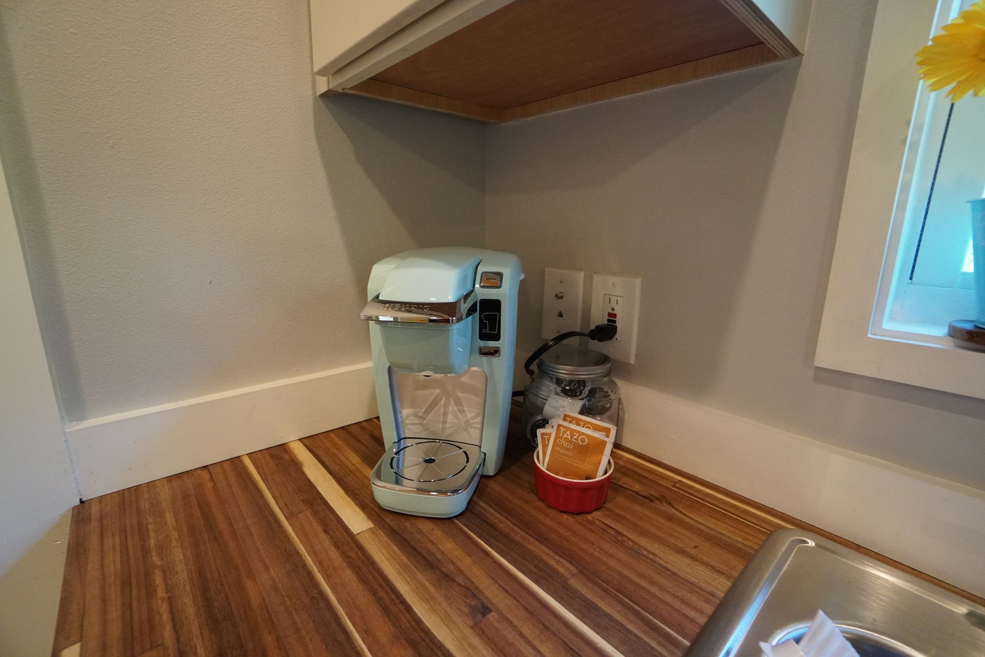 Coffee maker and tea accessories on a wooden countertop under a shelf. Light blue and red objects.