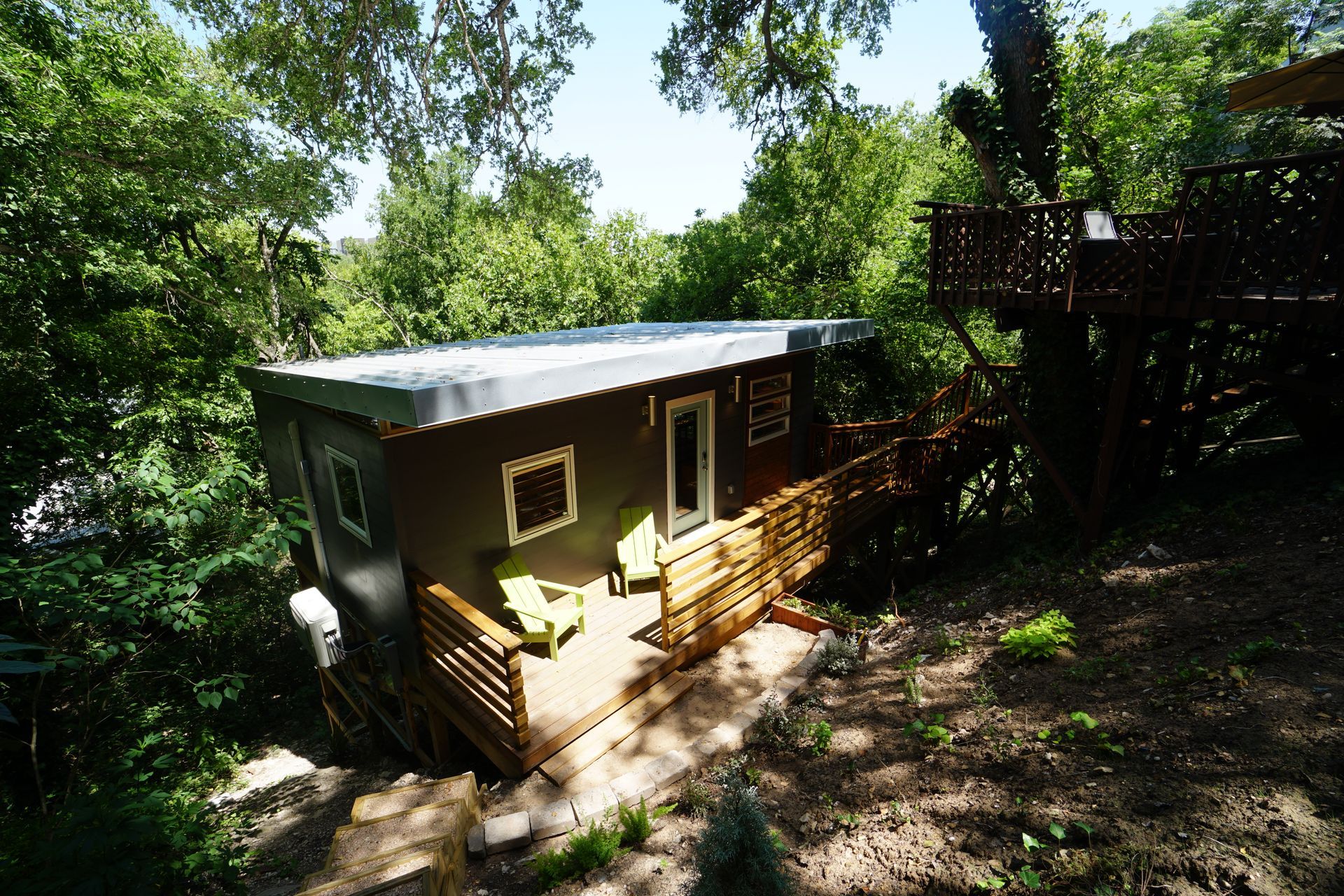 Cabin with a small deck surrounded by trees. Dark gray exterior, green chairs.