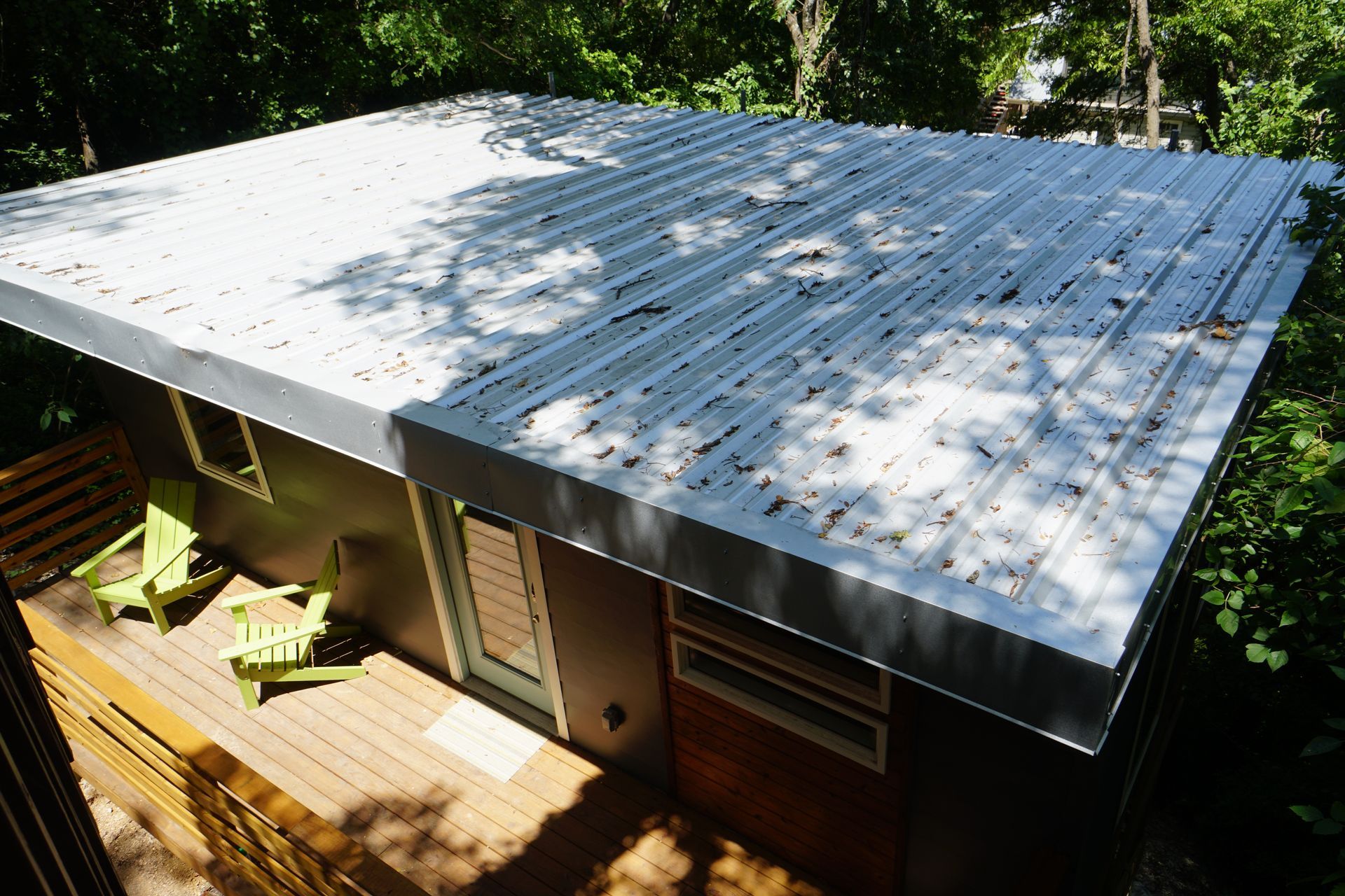 Modern house with a flat, corrugated metal roof and a wooden deck. Two green chairs sit on the deck.
