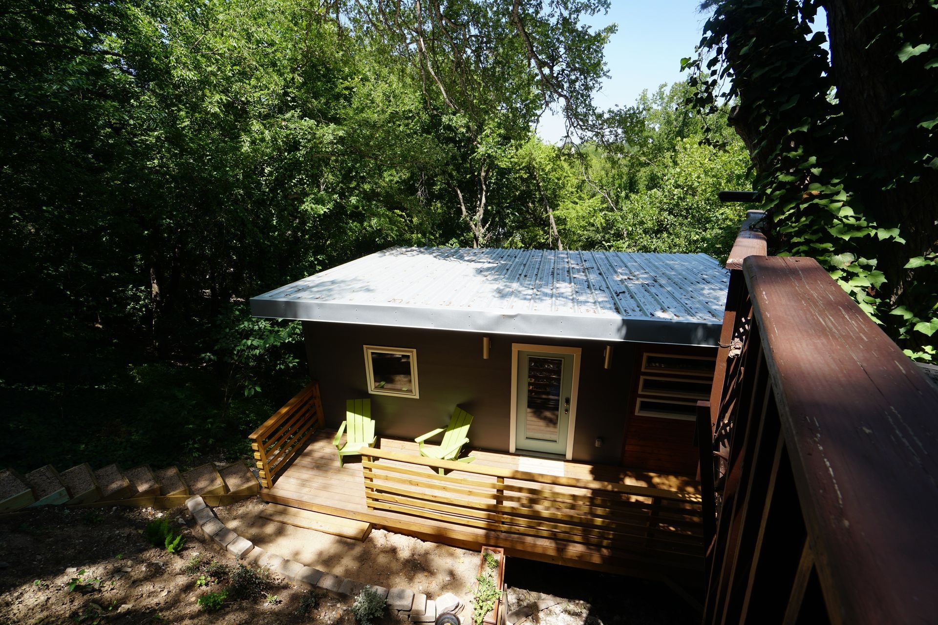 Small brown cabin with porch and metal roof in a wooded area; two green chairs on porch.