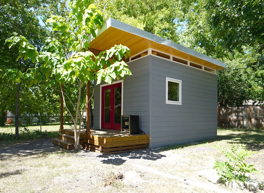 Gray, modern shed with a red door, small windows, and a wooden deck under trees.