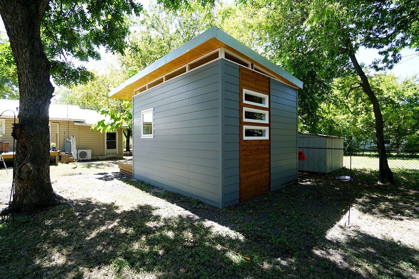 Modern gray and wood-paneled shed with small windows, sitting in a grassy yard under trees.