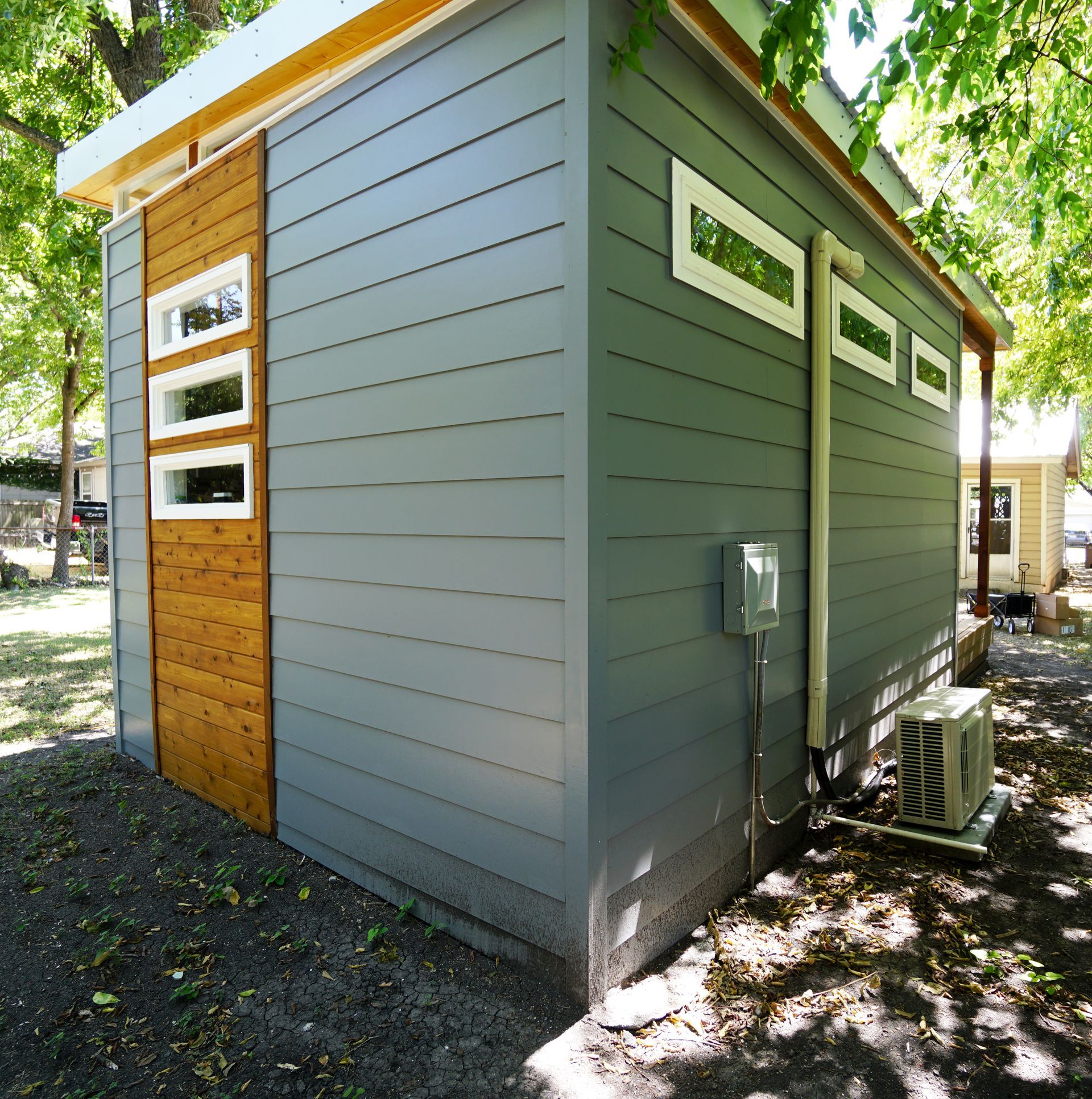 Modern gray and wood-sided shed with white-framed windows, outdoor unit, and small electrical box.