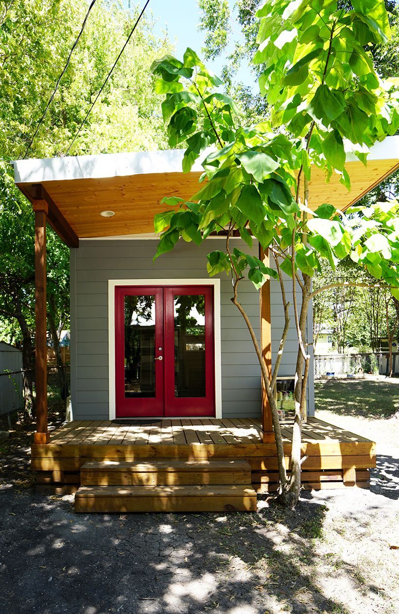 Gray building with red double doors, wooden porch, and canopy, surrounded by trees.