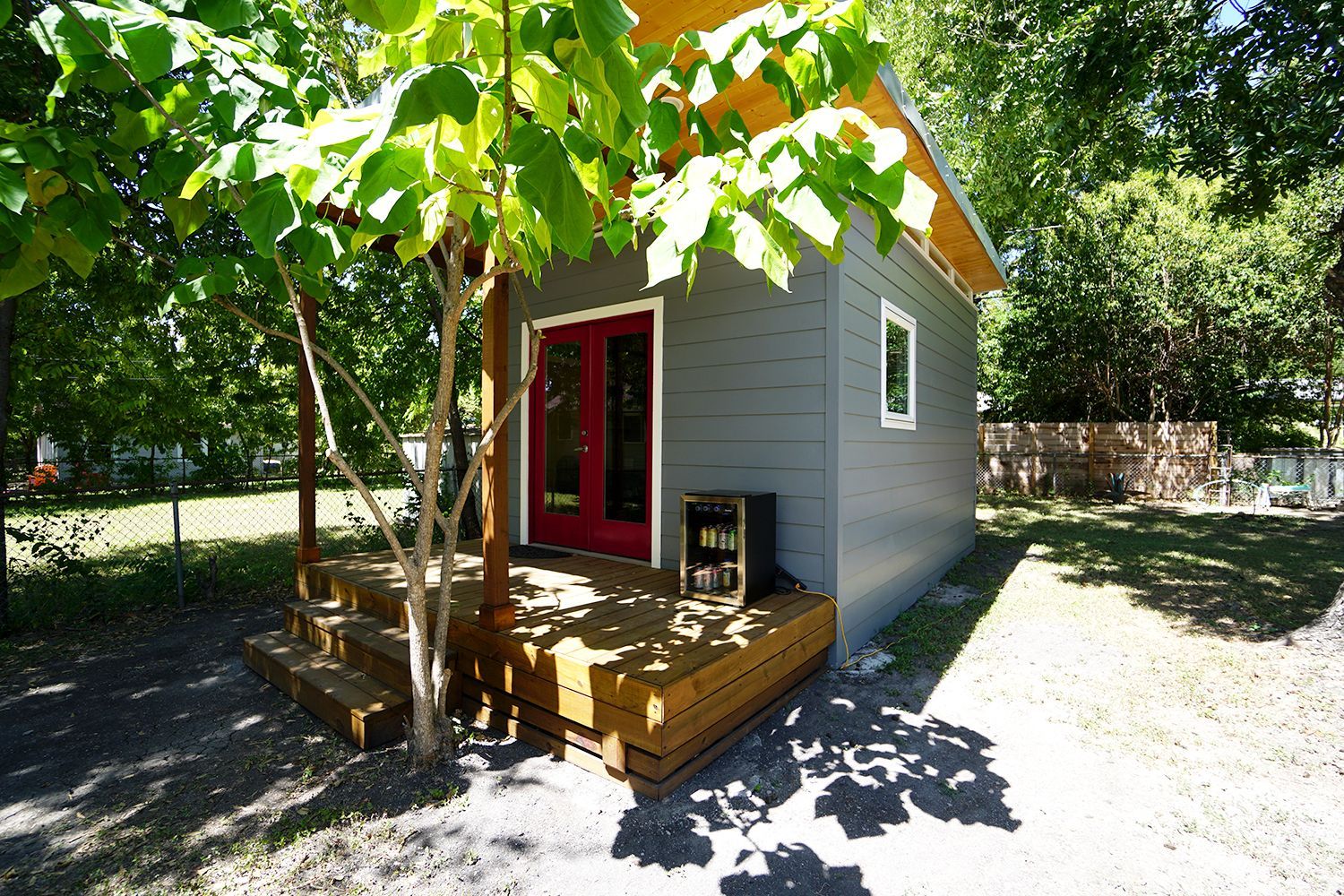 Small gray cabin with red double doors and a wooden deck, shaded by a tree.