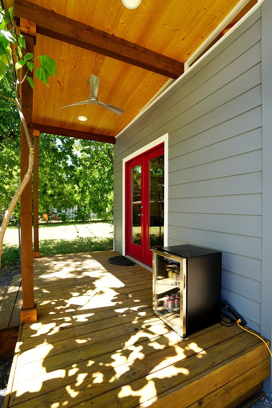 Covered porch with gray siding, red door, and a mini-fridge. Wooden beams and ceiling. Sunny day.