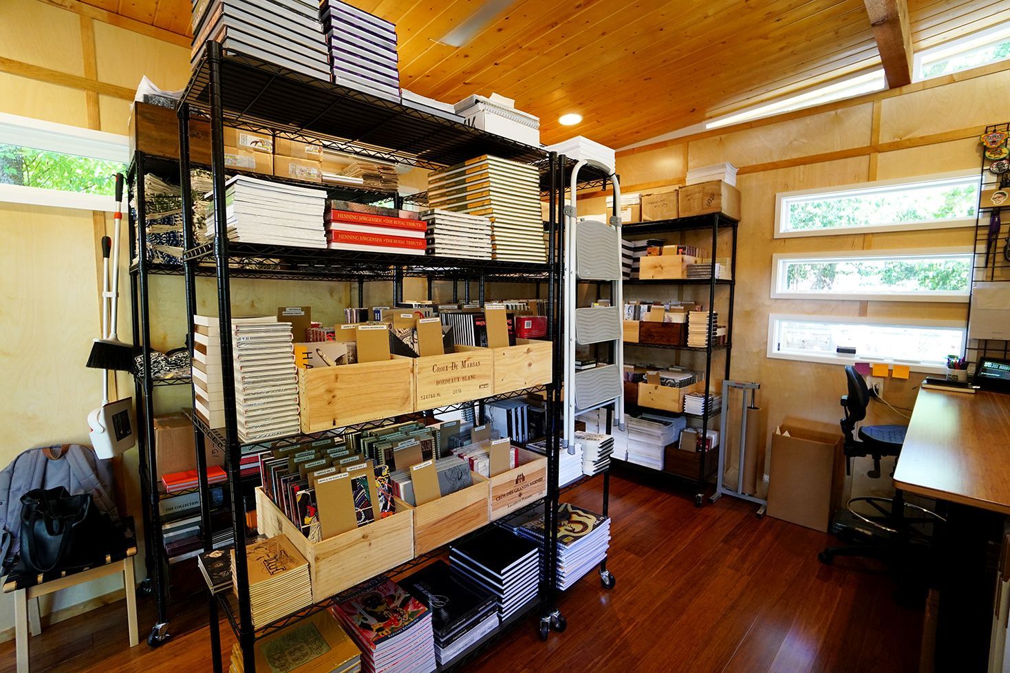 Shelves in a cluttered storage room holding boxes and crates of goods, with a desk and windows in the background.