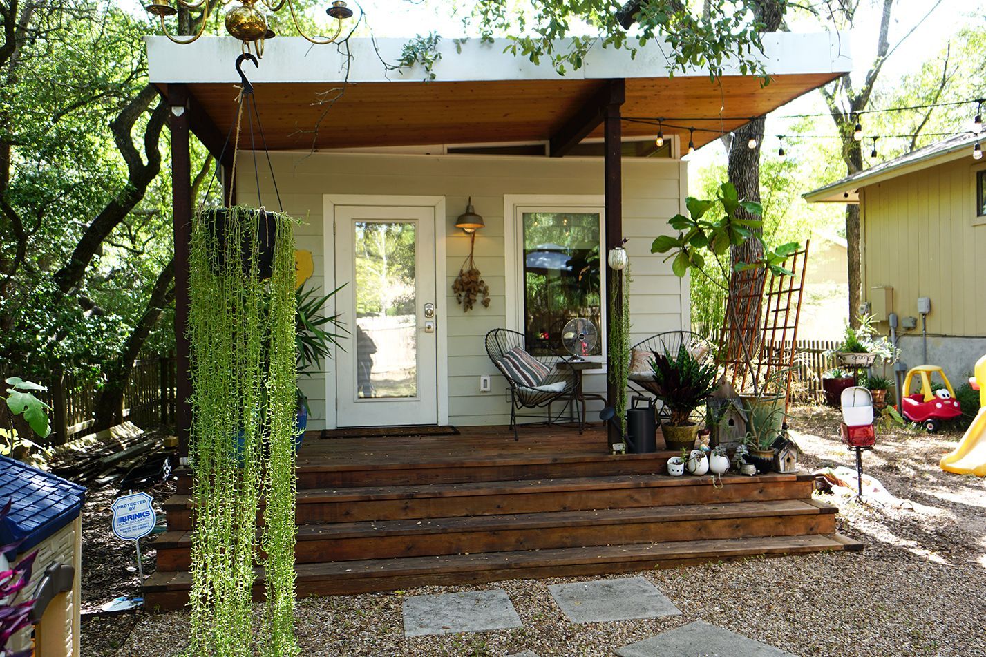 A small white outdoor building with a wooden porch and steps, surrounded by greenery.
