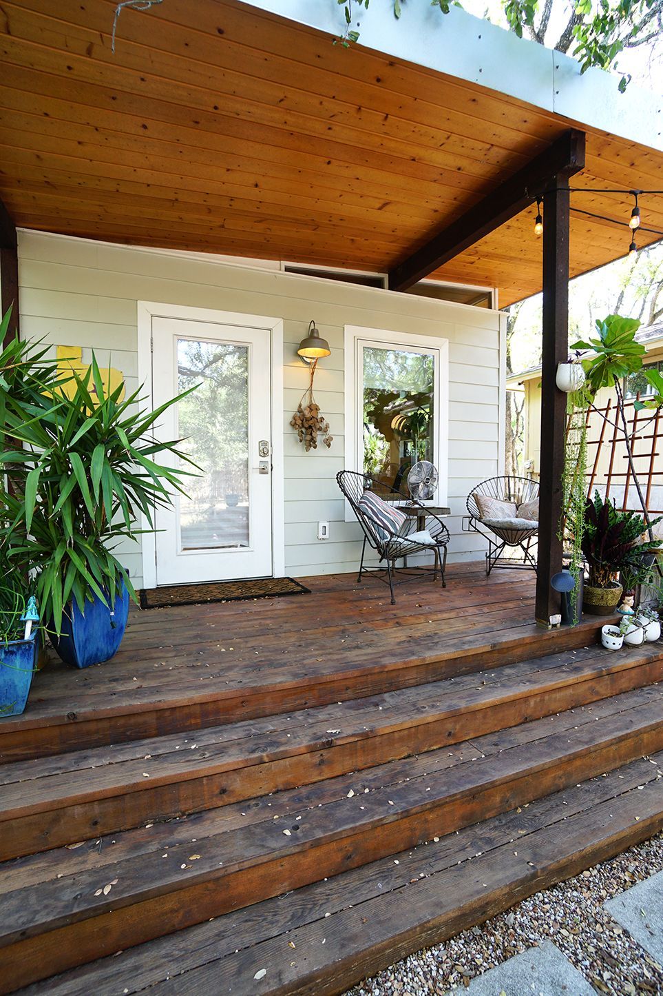 Wooden deck with steps leading up to a light green house with a porch, chairs, and plants.
