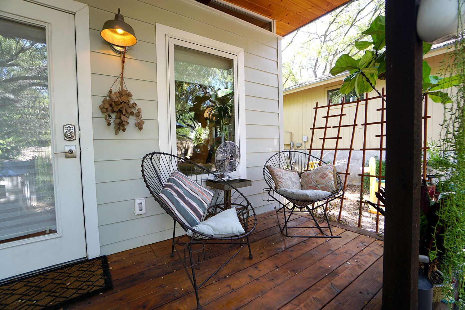 Cozy porch with two wrought iron chairs, small table, and a window. Wood deck and green foliage.