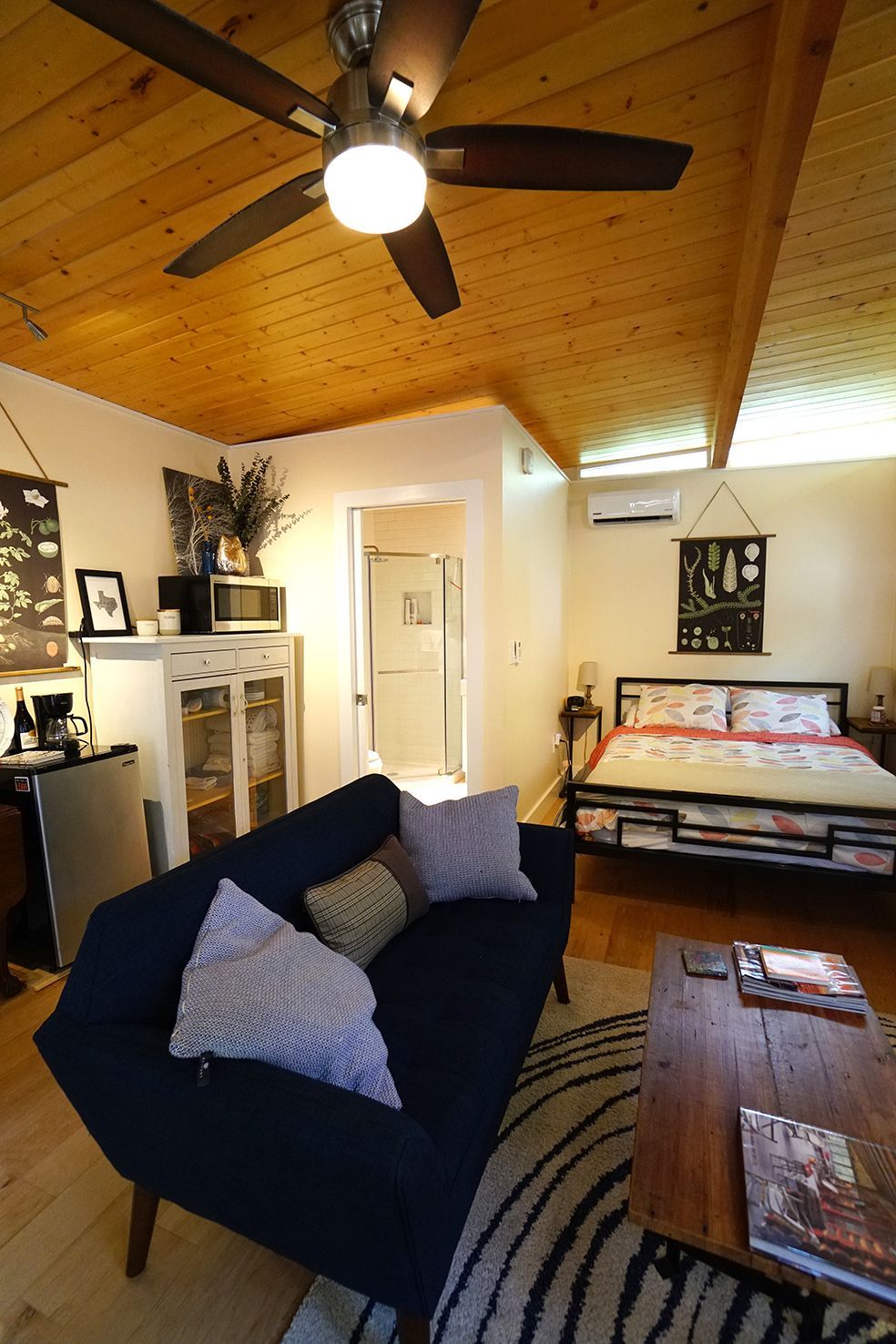Small cabin interior with blue couch, bed, wood ceiling, and round rug.