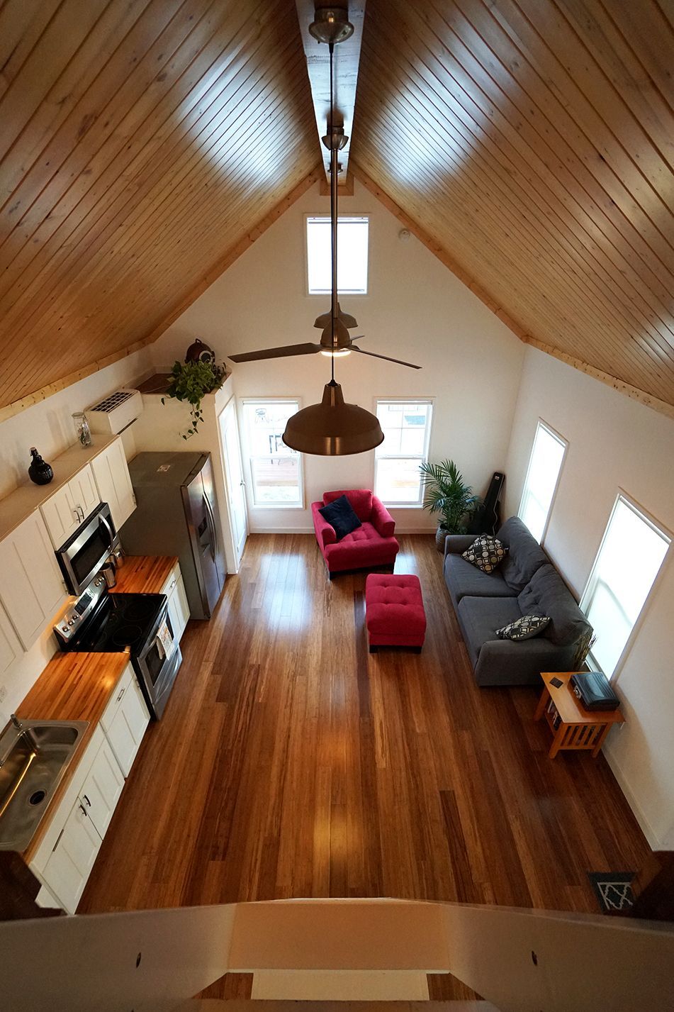 A wide shot of a living space with a kitchen. Hardwood floors and wood-paneled ceiling. Windows and seating are visible.