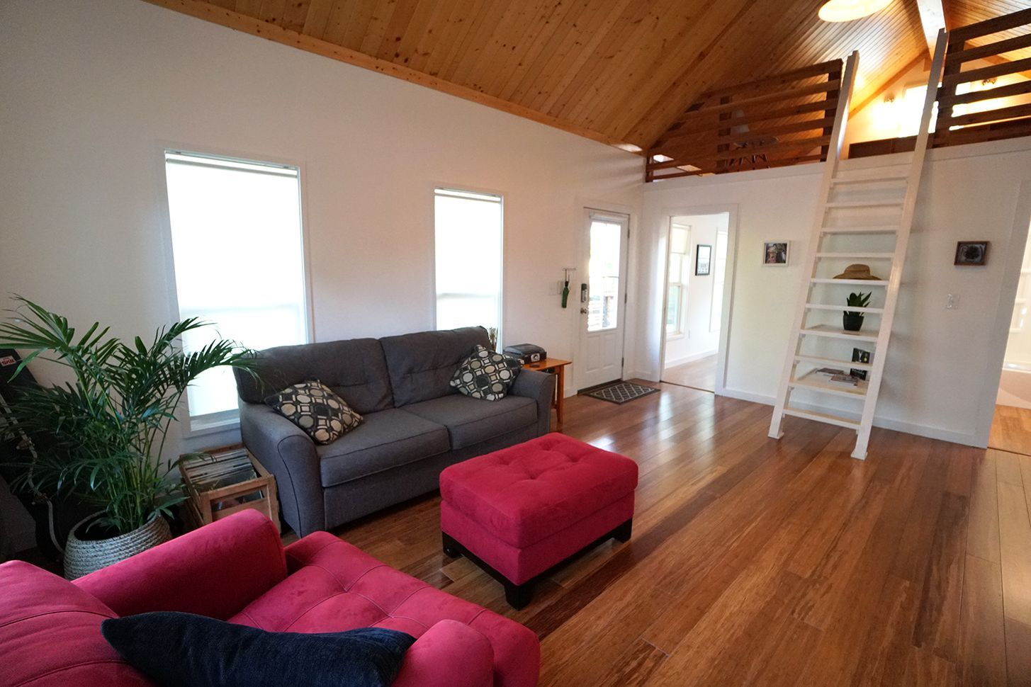Living room with wood floors, two sofas, ottoman, ladder to loft, and large windows.