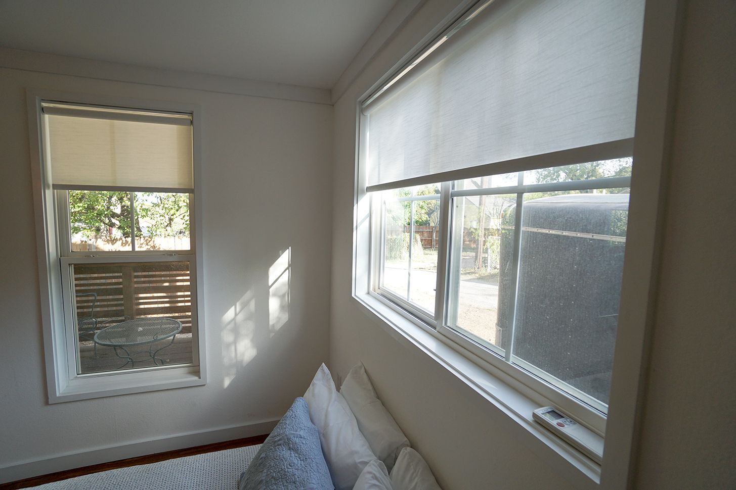 Two windows with white frames and beige roller shades; a glimpse of a backyard is seen through the glass.
