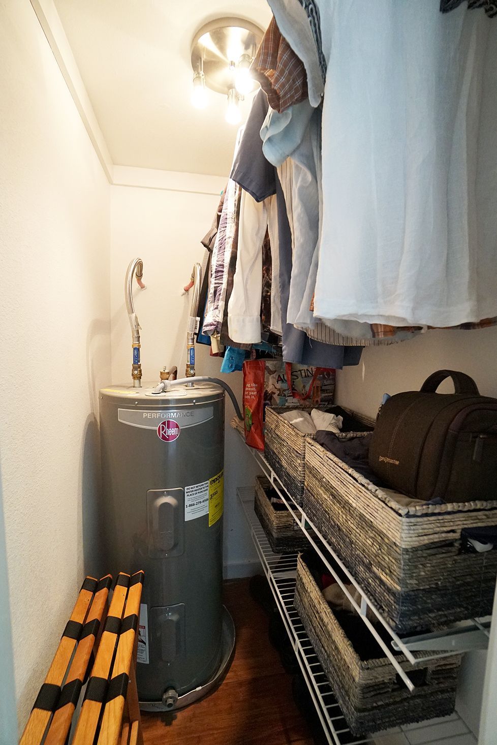 A closet with a water heater, clothes on a rack, and shelves.